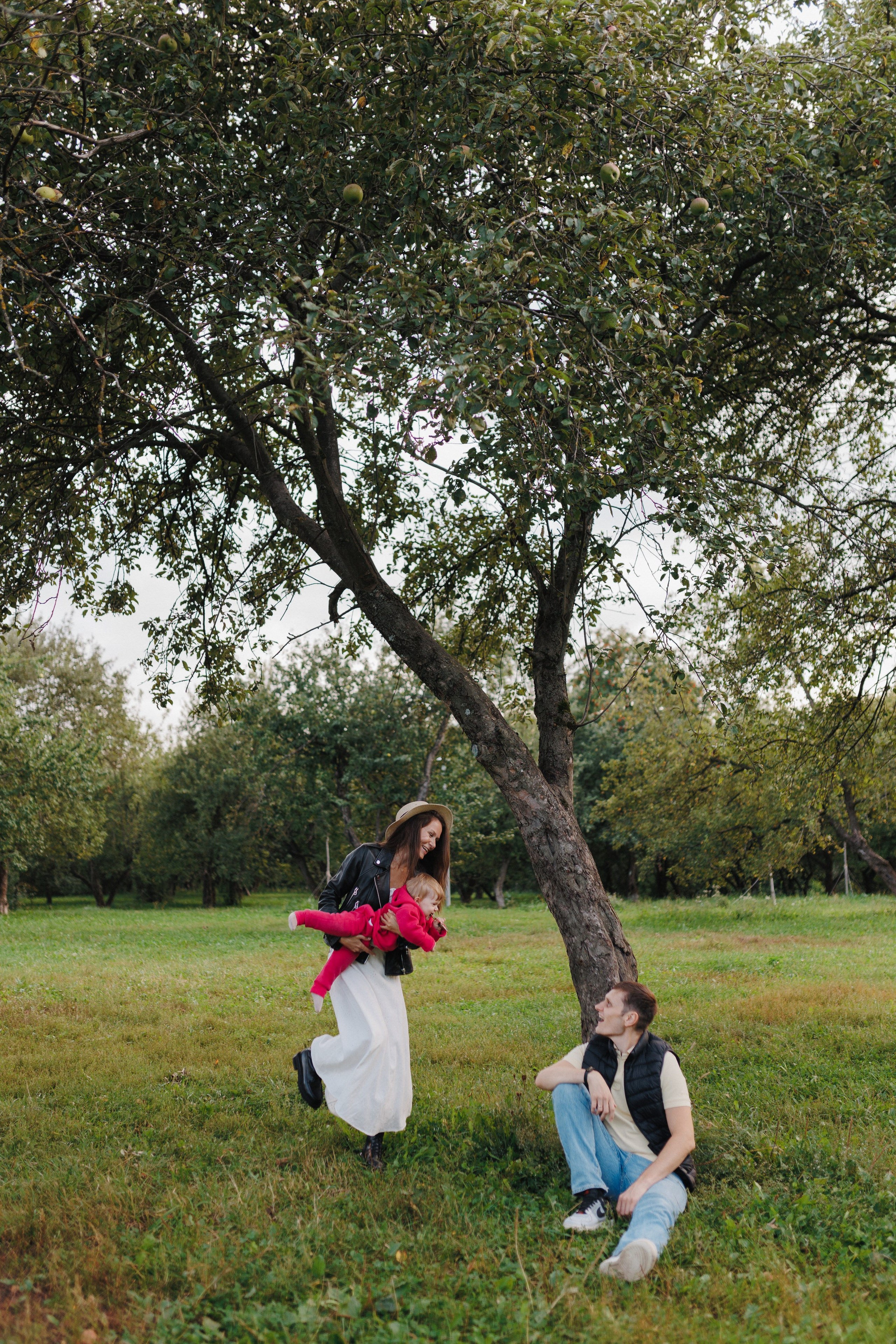 Отец сидит под деревом, а мама рядом держит на руках их общего ребёнка. Естественные и нежные семейные кадры.