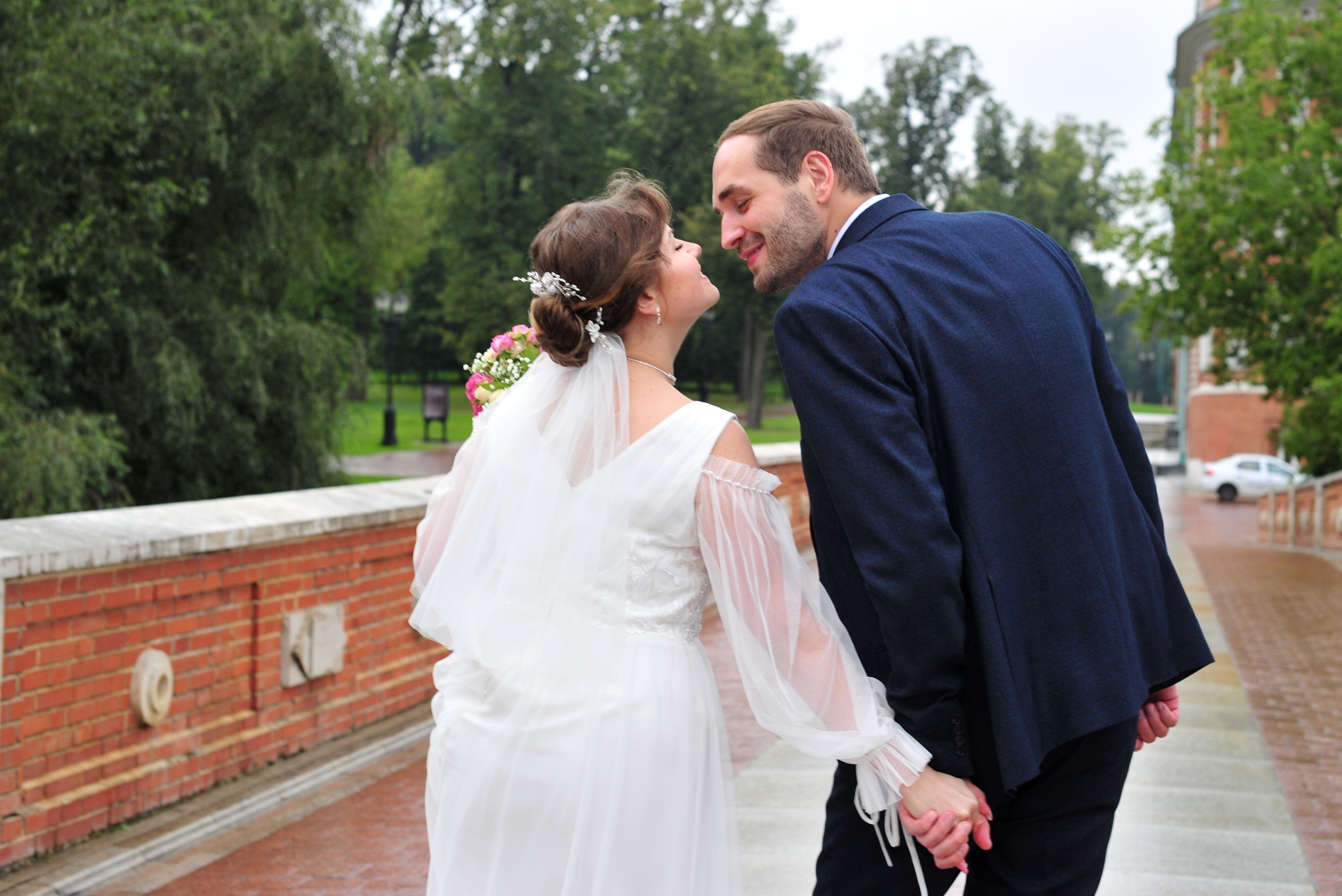 Свадьба в Царицыно пасмурным летним днем, поцелуй новобрачных, вид со спины жениха и невесты на мосту из красного кирпича и белого камня 