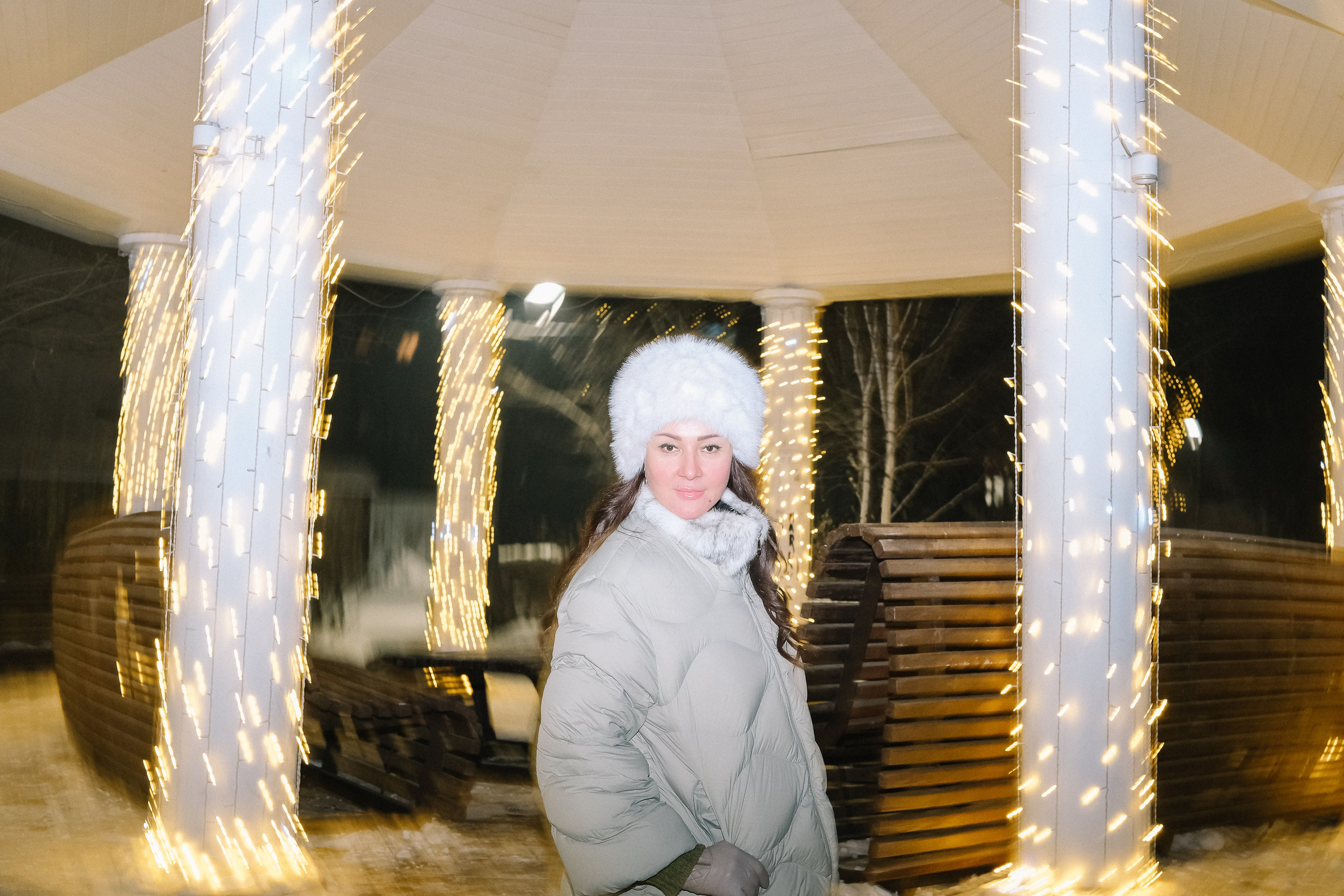 Новогодняя фотосессия: женщина в светлом пуховике среди сияющих колонн беседки