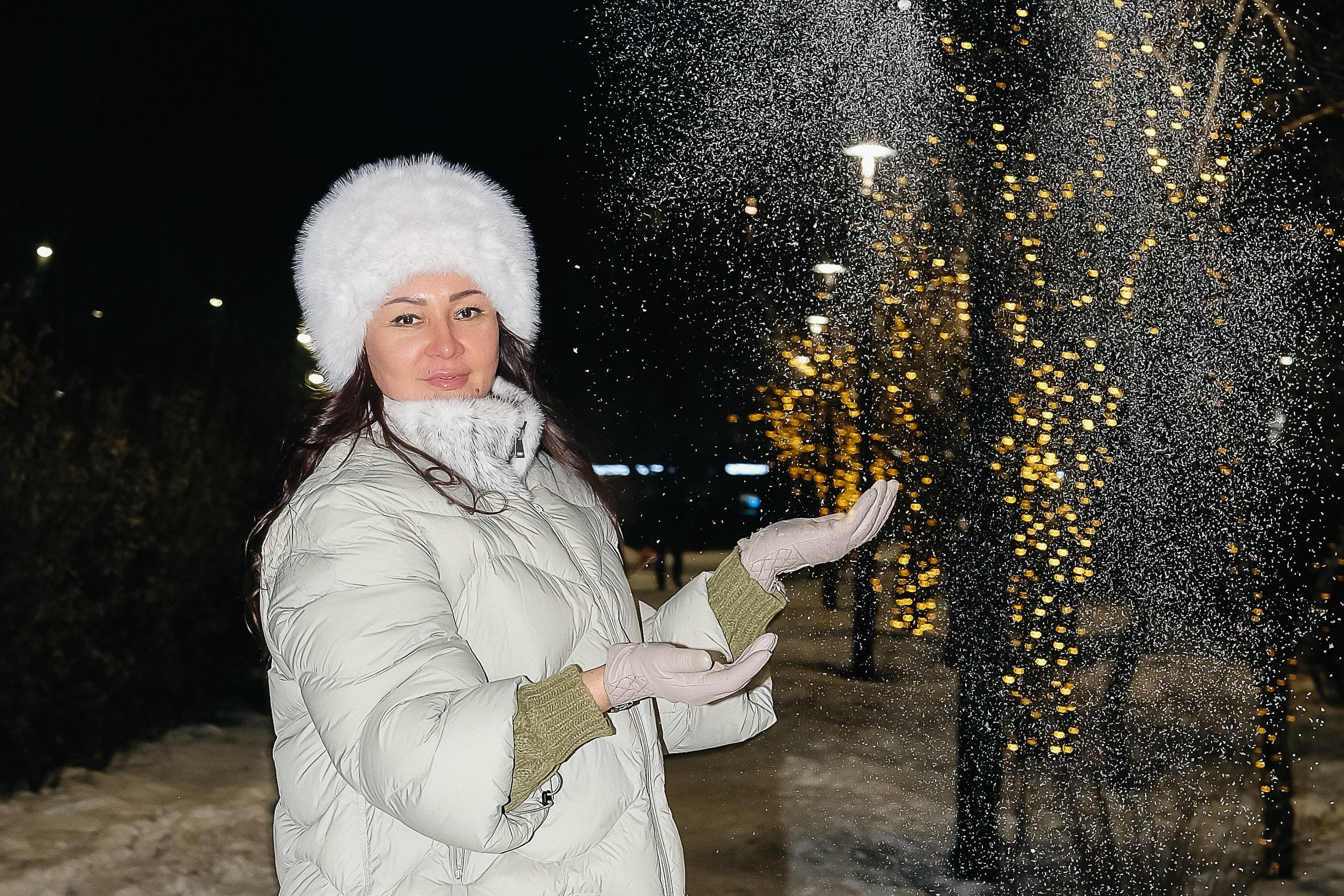 Женщина подбрасывает снег на вечерней прогулке среди праздничных огней
