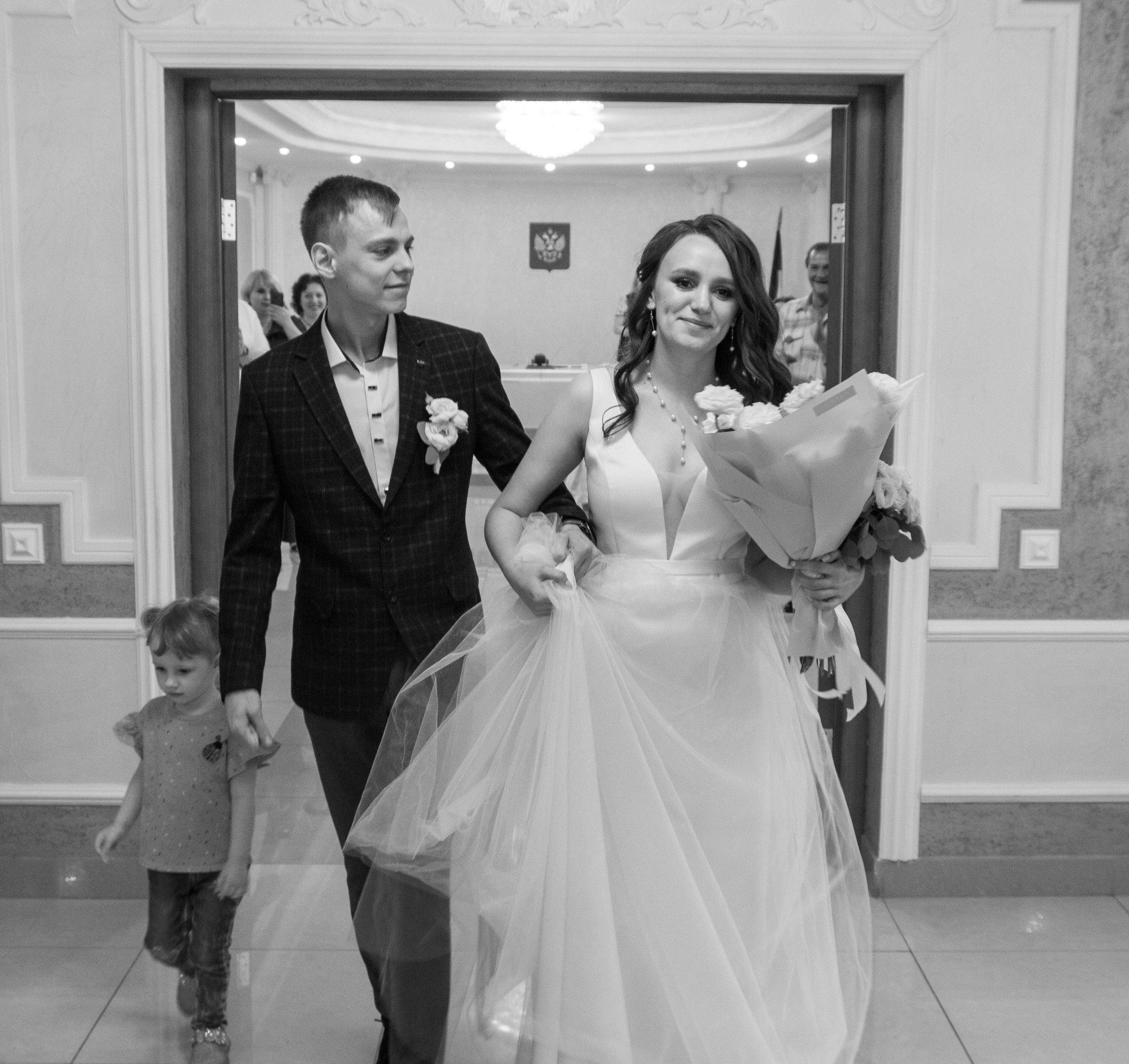 Чёрно-белое фото — невеста идёт с букетом и улыбается, рядом идут жених и ребёнок.