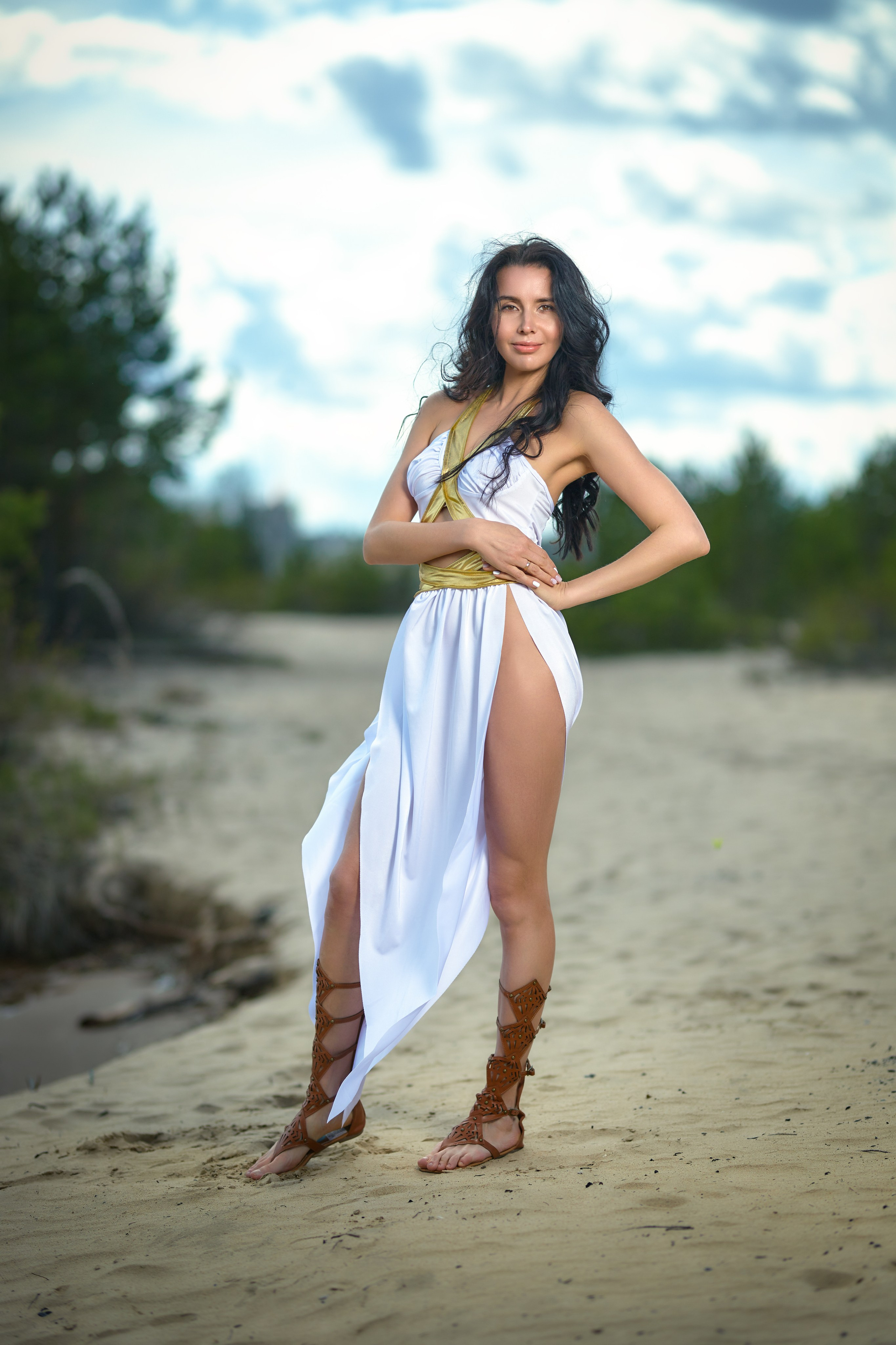 Девушка в белой греческой одежде и кожаных сандалях стоит на песке