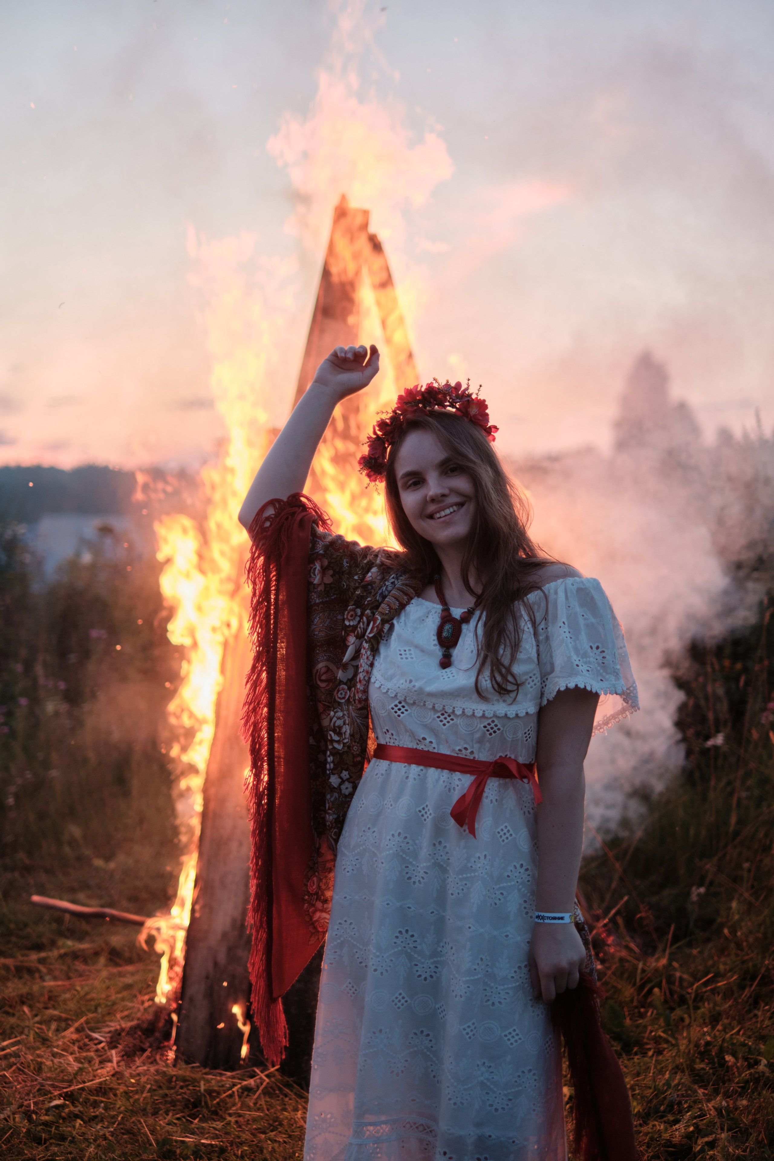 Девушка стоит в русском костюме и венком сзади горит большой костер