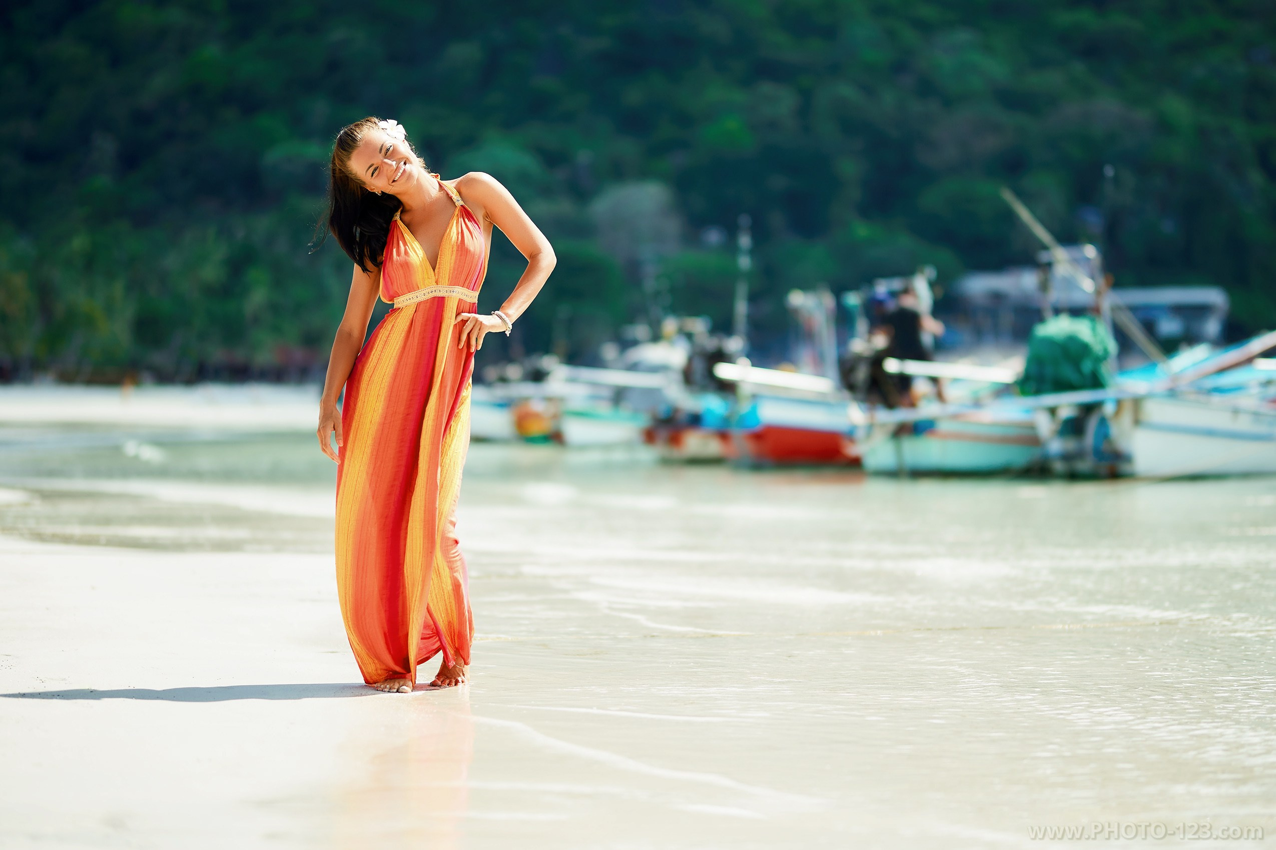 Woman in a flowing orange dress standing barefoot on wet sand near fishing boats, soft sunlight, calm tropical beach atmosphere, photographed on Phu Quoc by a local photographer