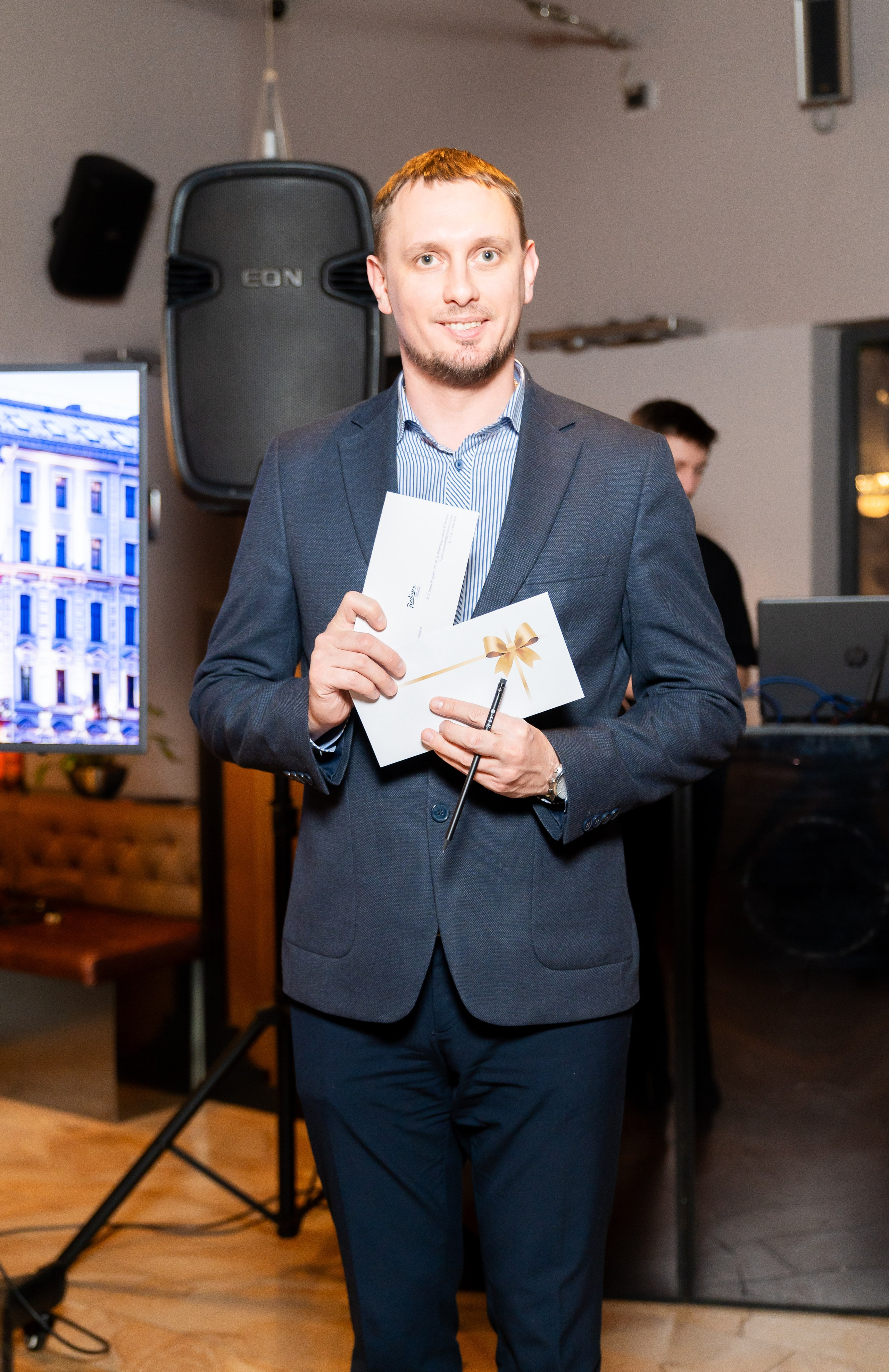 Мужчина в синем костюме с конвертом и подарочным сертификатом Radisson позирует на корпоративном мероприятии. Фотография Евгения Мальцева.