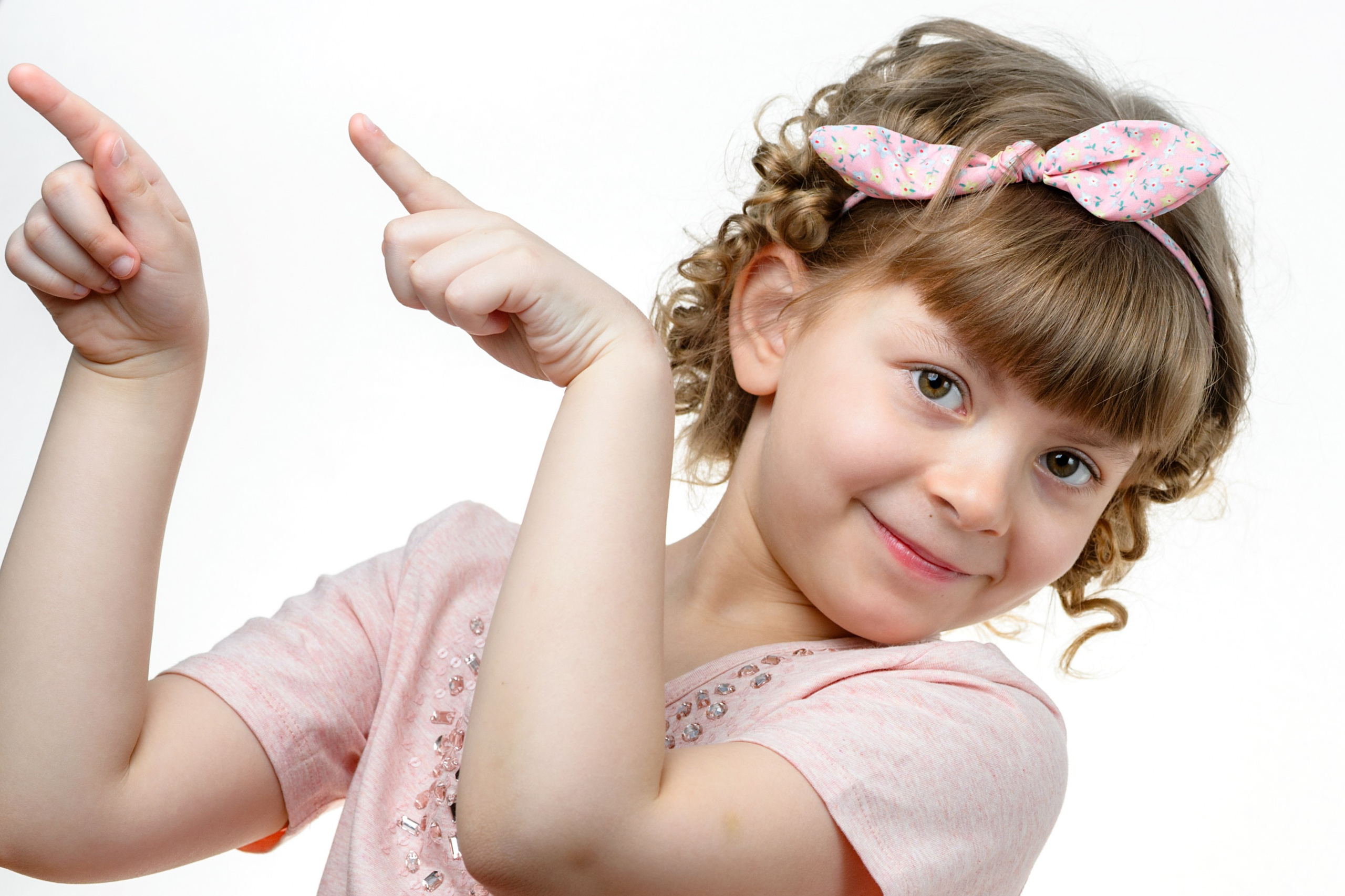 Девочка с бантиками наклоняется к камере и указывает рукой в сторону на белом фоне.