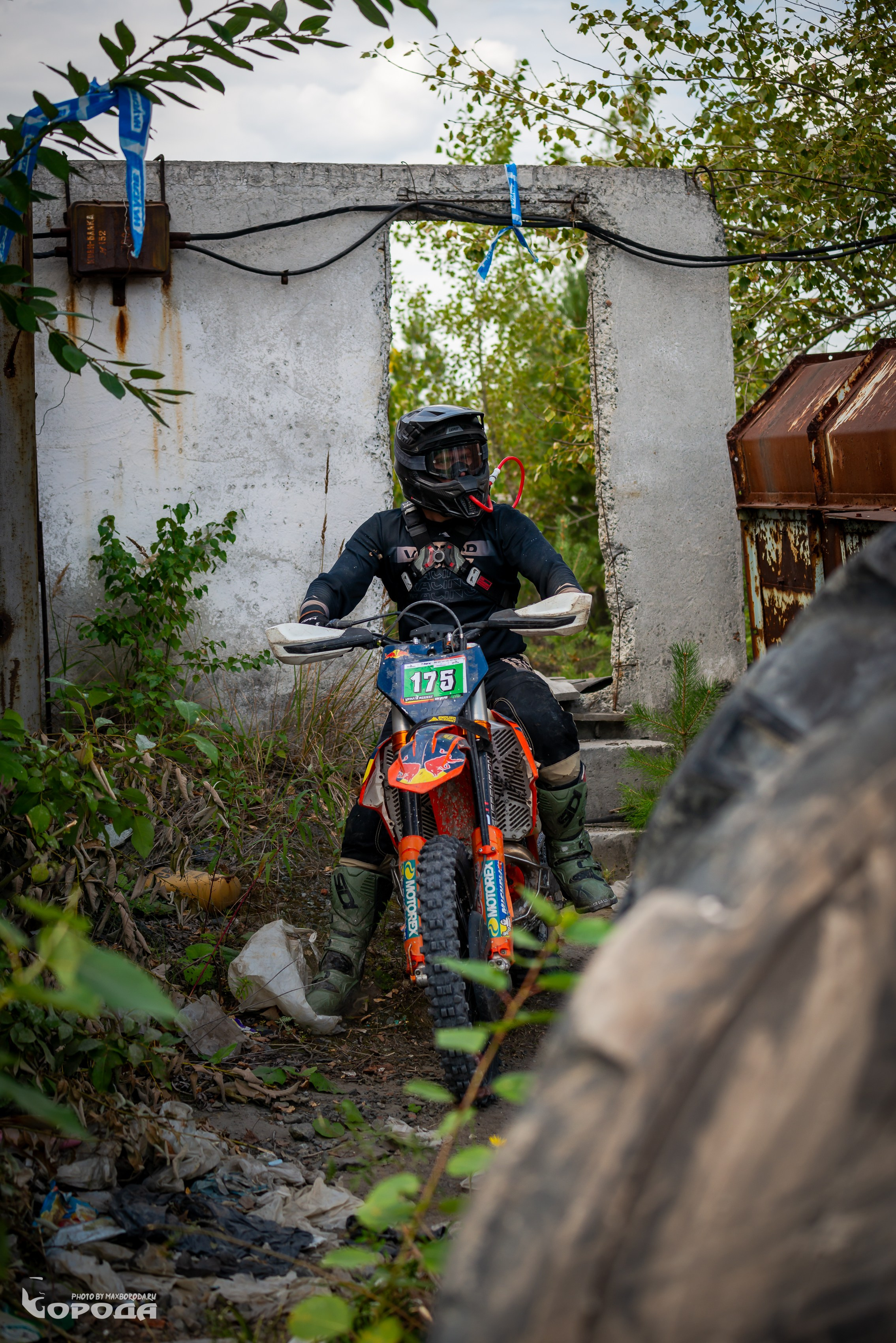 Гонщик на эндуро возле ржавых конструкций и зелёных кустов.