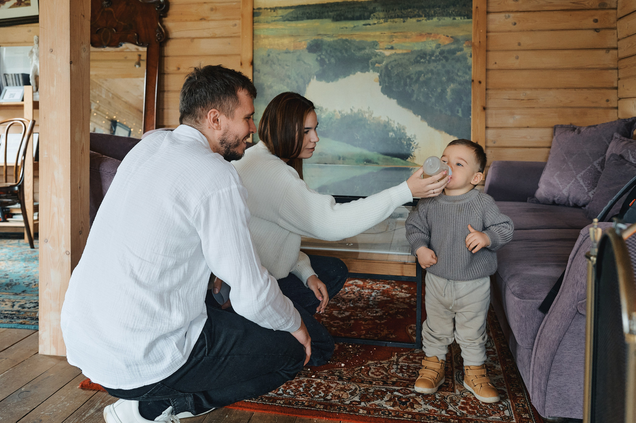 семья в интерьере: мама поит ребёнка из бутылочки, папа сидит рядом, уютная обстановка