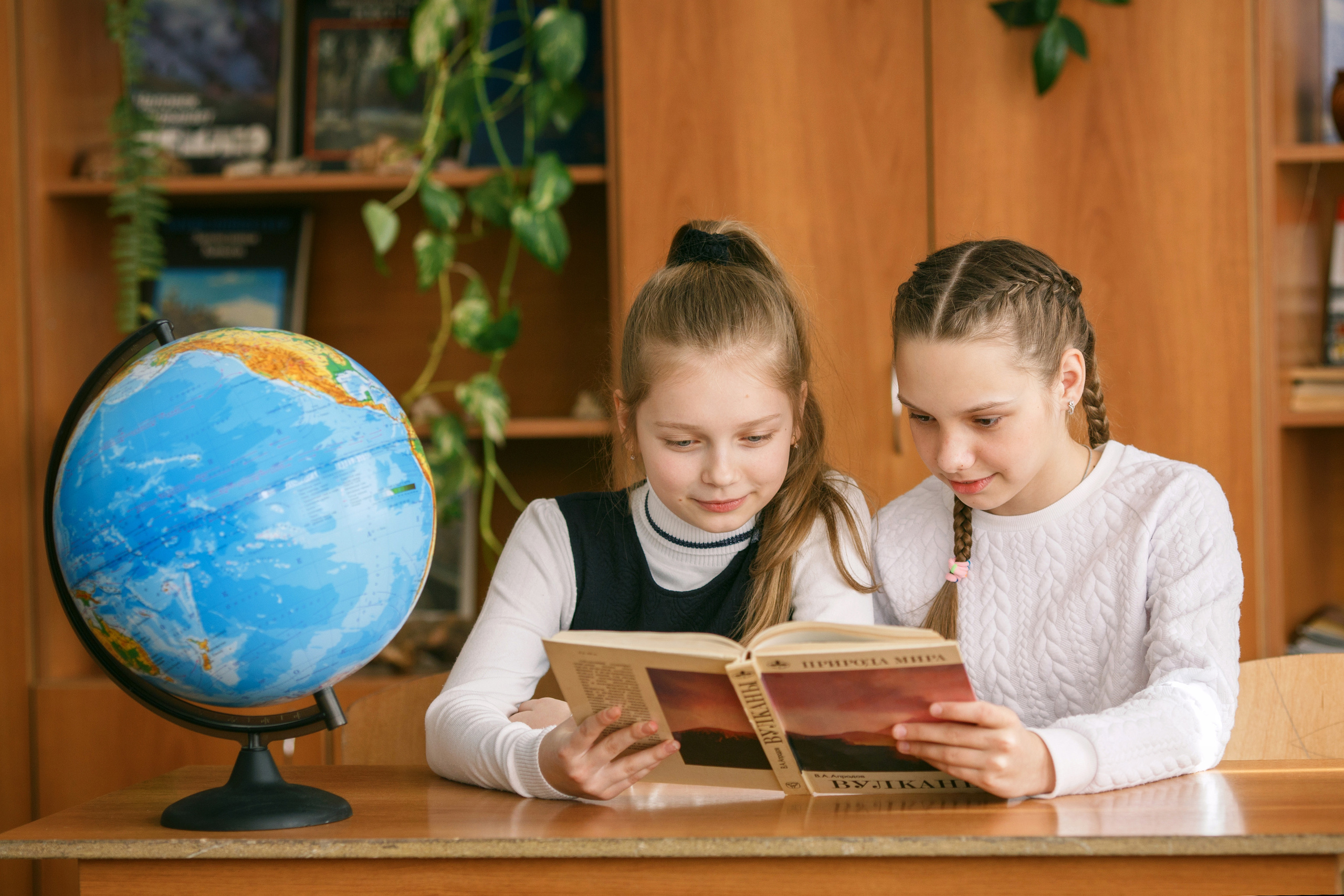Две девочки читают книгу за партой, на которой стоит глобус