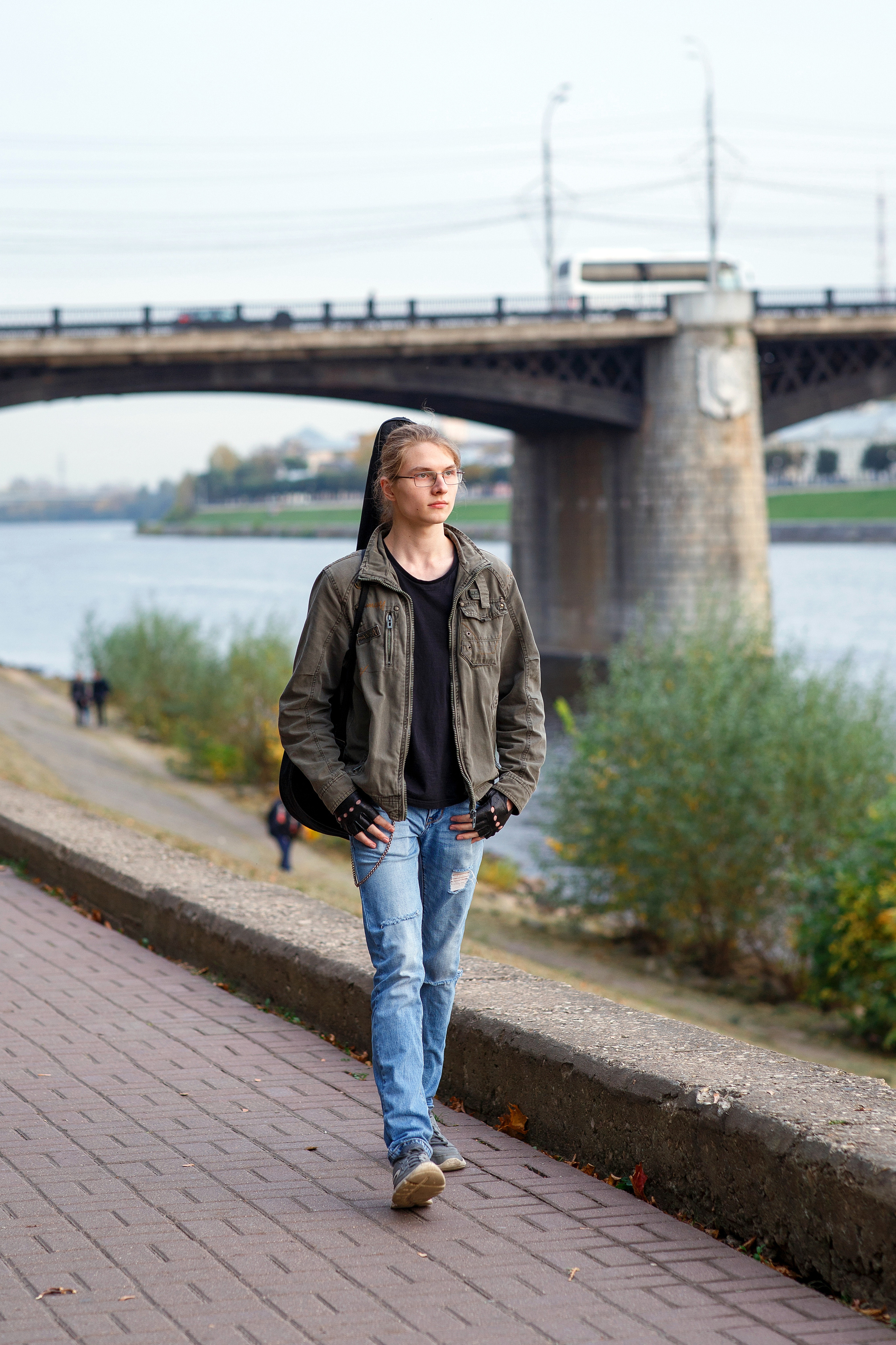 Парень в очках с гитарой в чехле идет по набережной