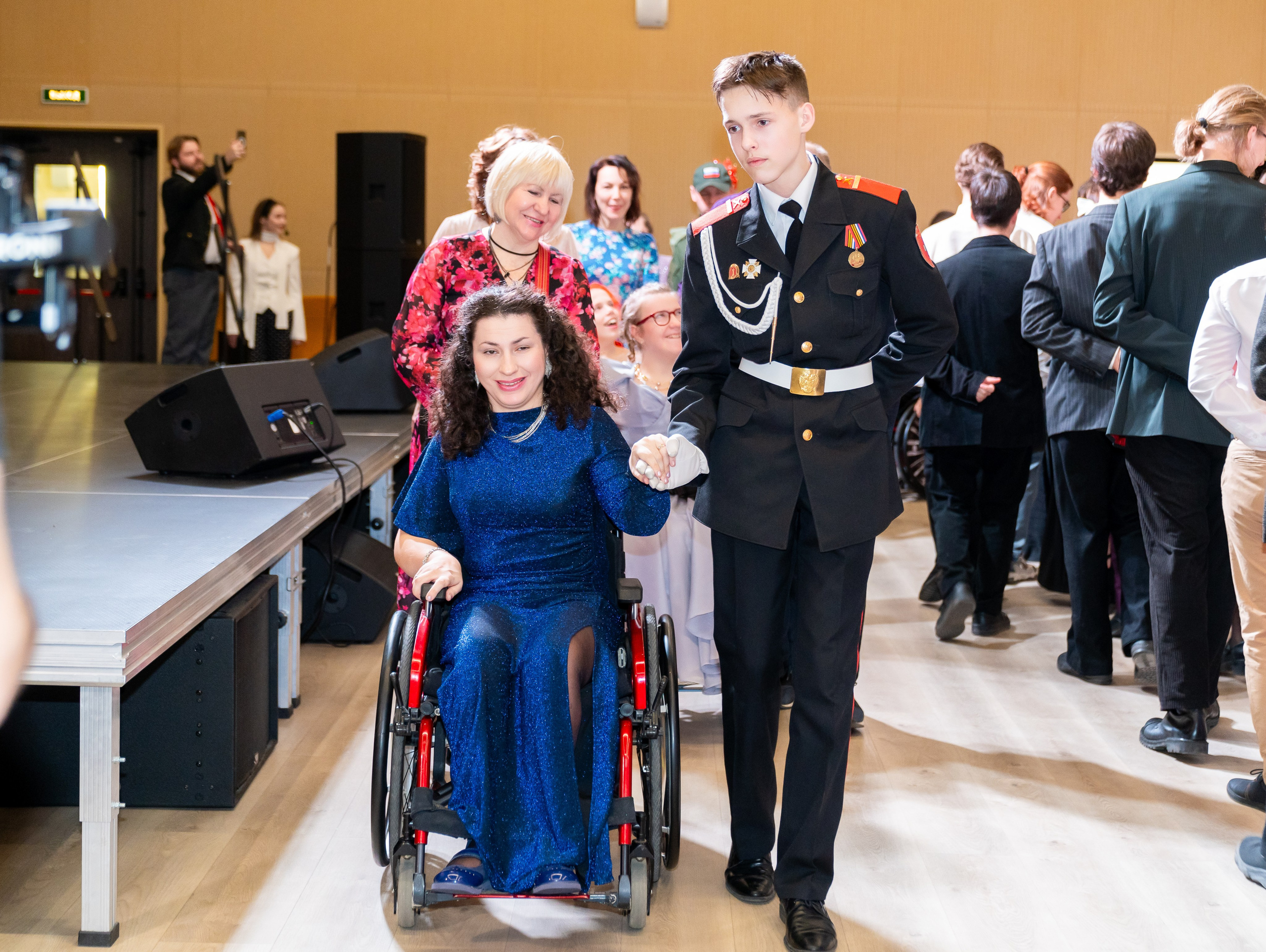 Юноша в парадной военной форме ведёт под руку женщину в синем блестящем платье на инвалидной коляске во время инклюзивного бала. Фотограф Мальцев Евгений.