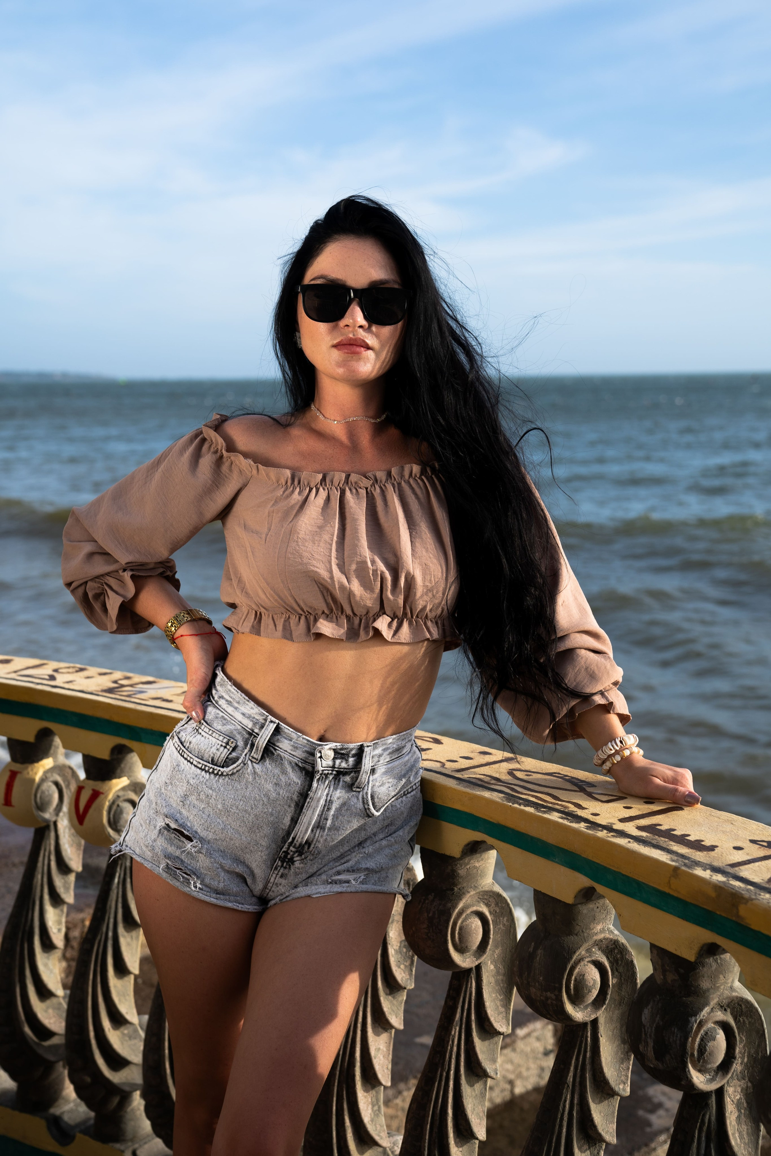 Девушка в шортах и топе позирует у каменного ограждения на фоне моря и неба.