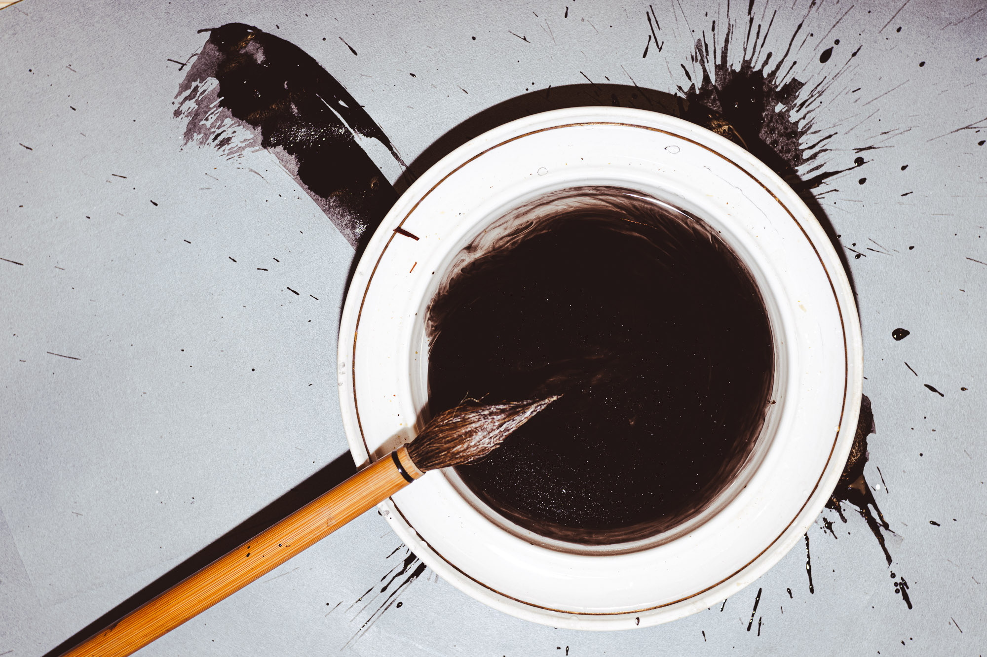 Вид сверху. В белом блюдце черные чернила, кисть для каллиграфии окунается в чернила. Серый фон