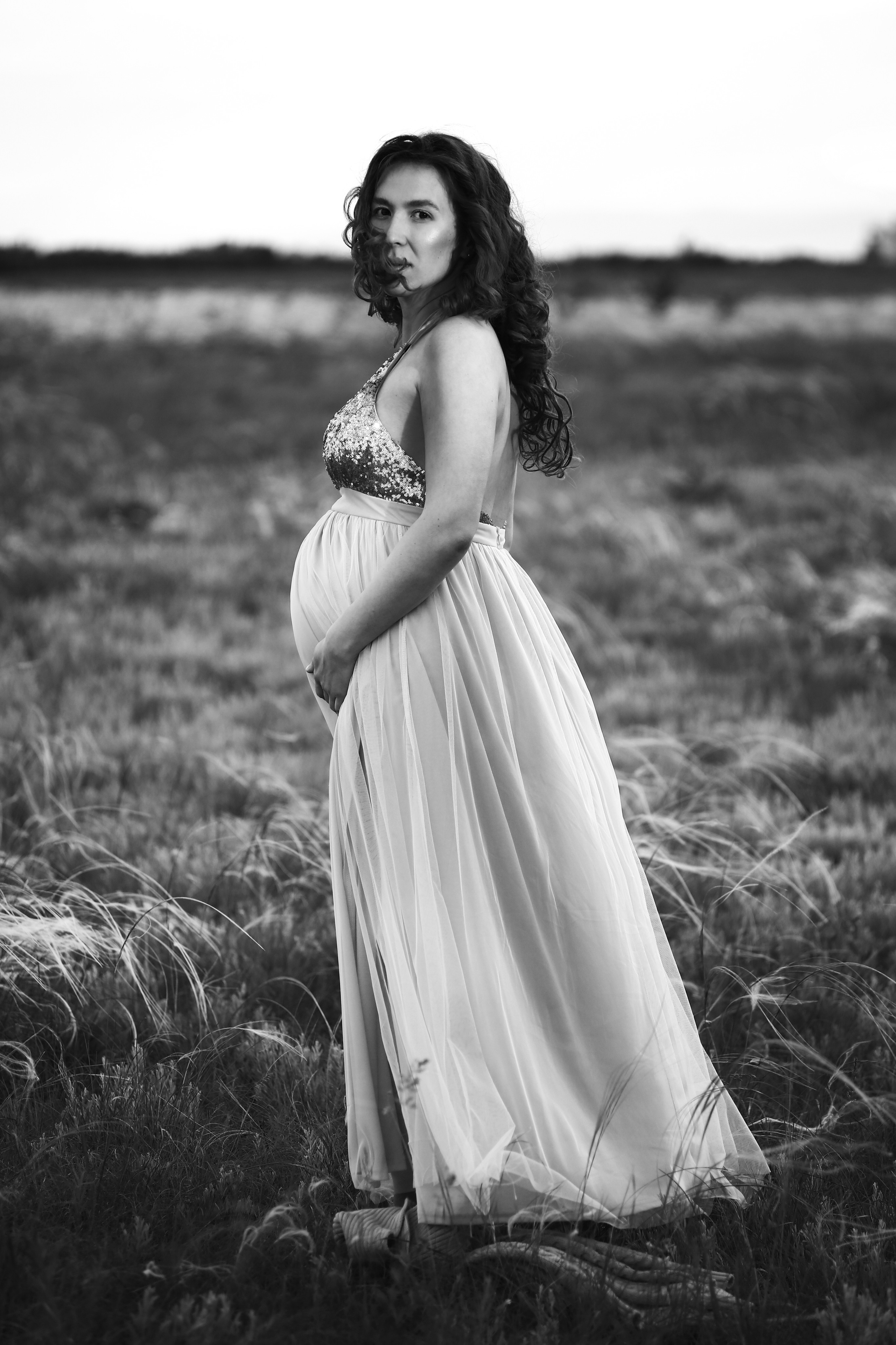 Беременная девушка в поле стоит в лучах заката в красивом длинном платье