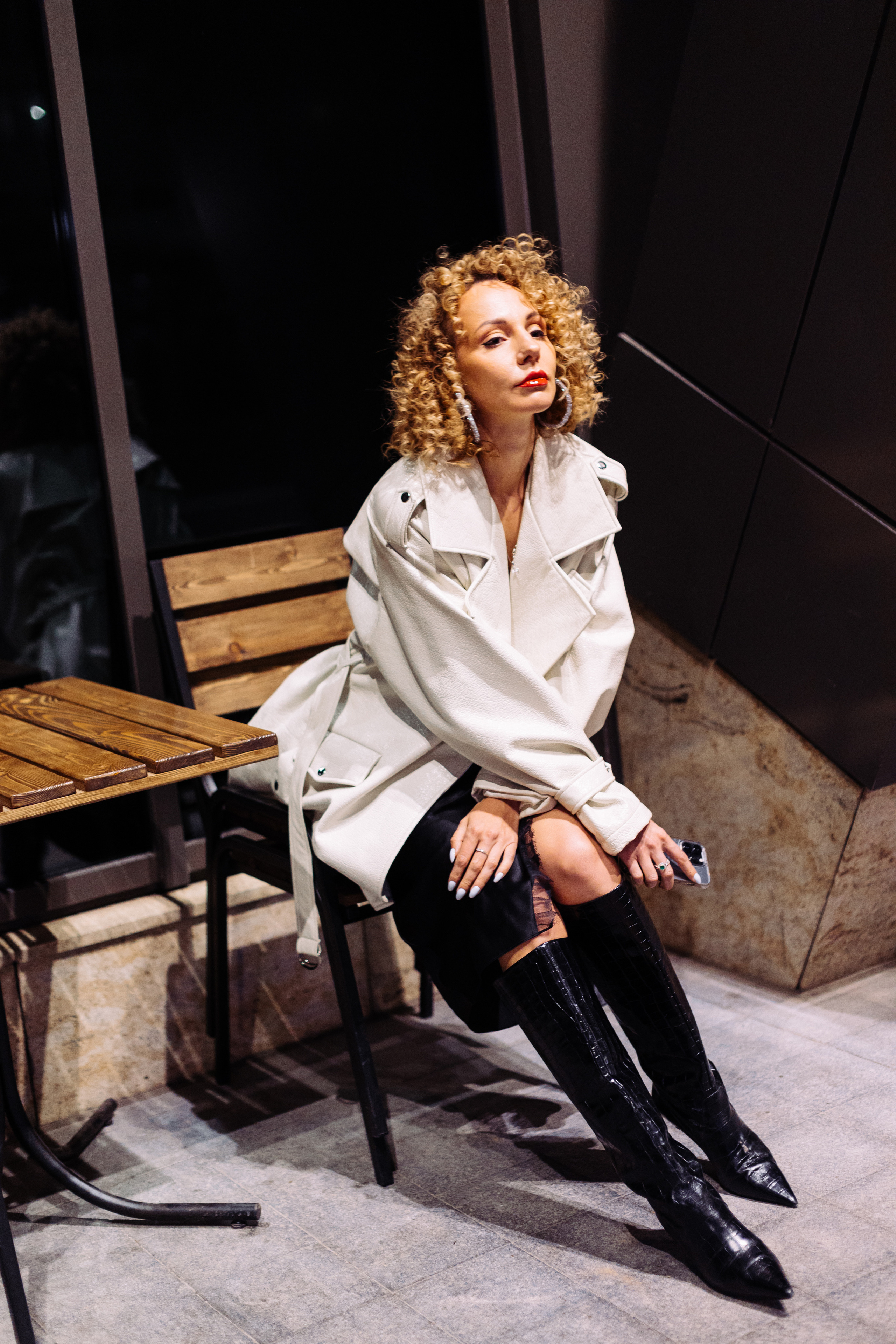 девушка сидящая за столиком кафе в ночном городе в белом плаще и красивом бельевом платье 