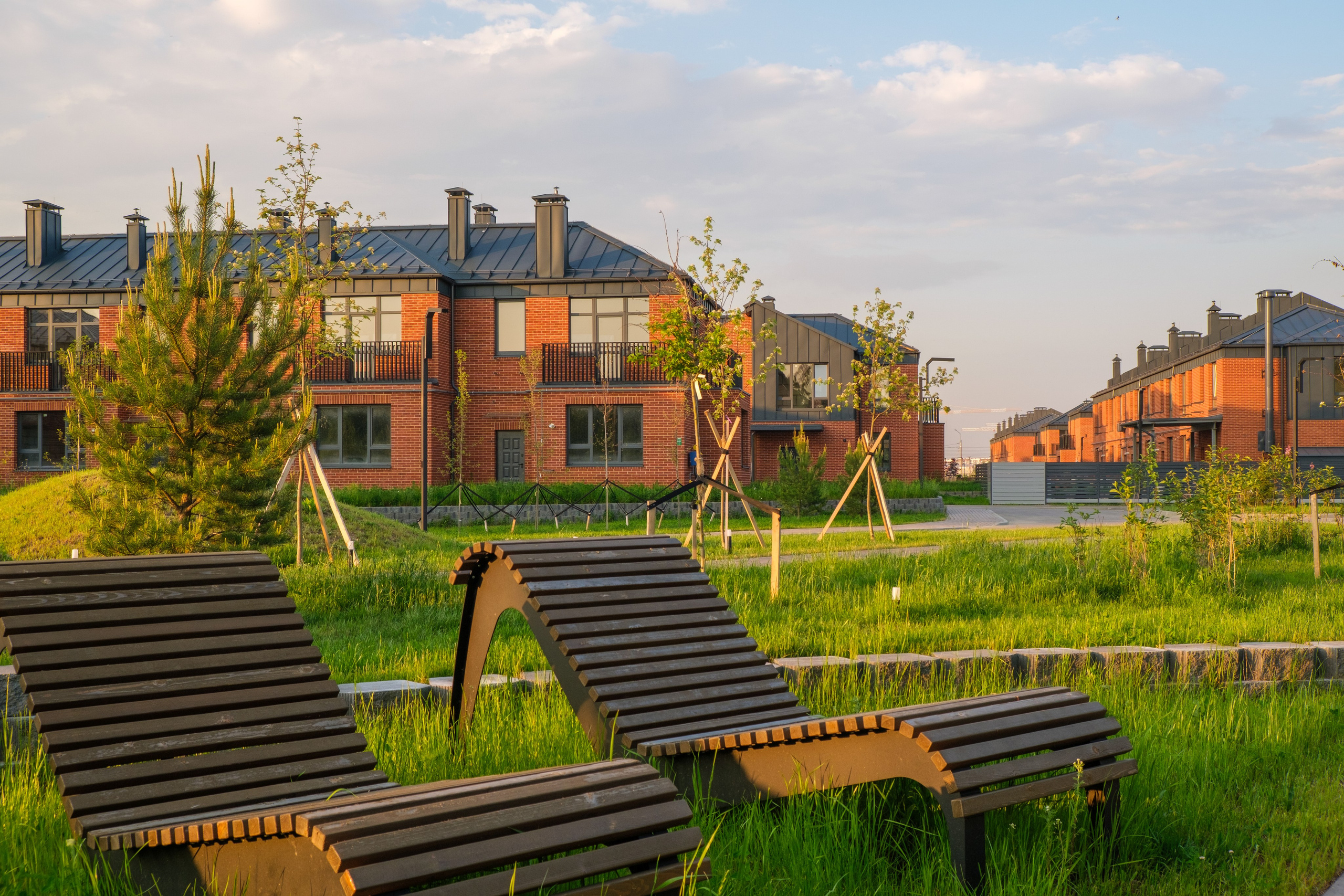 Две деревянные скамейки на переднем плане. На фоне - современный коттеджный посёлок.