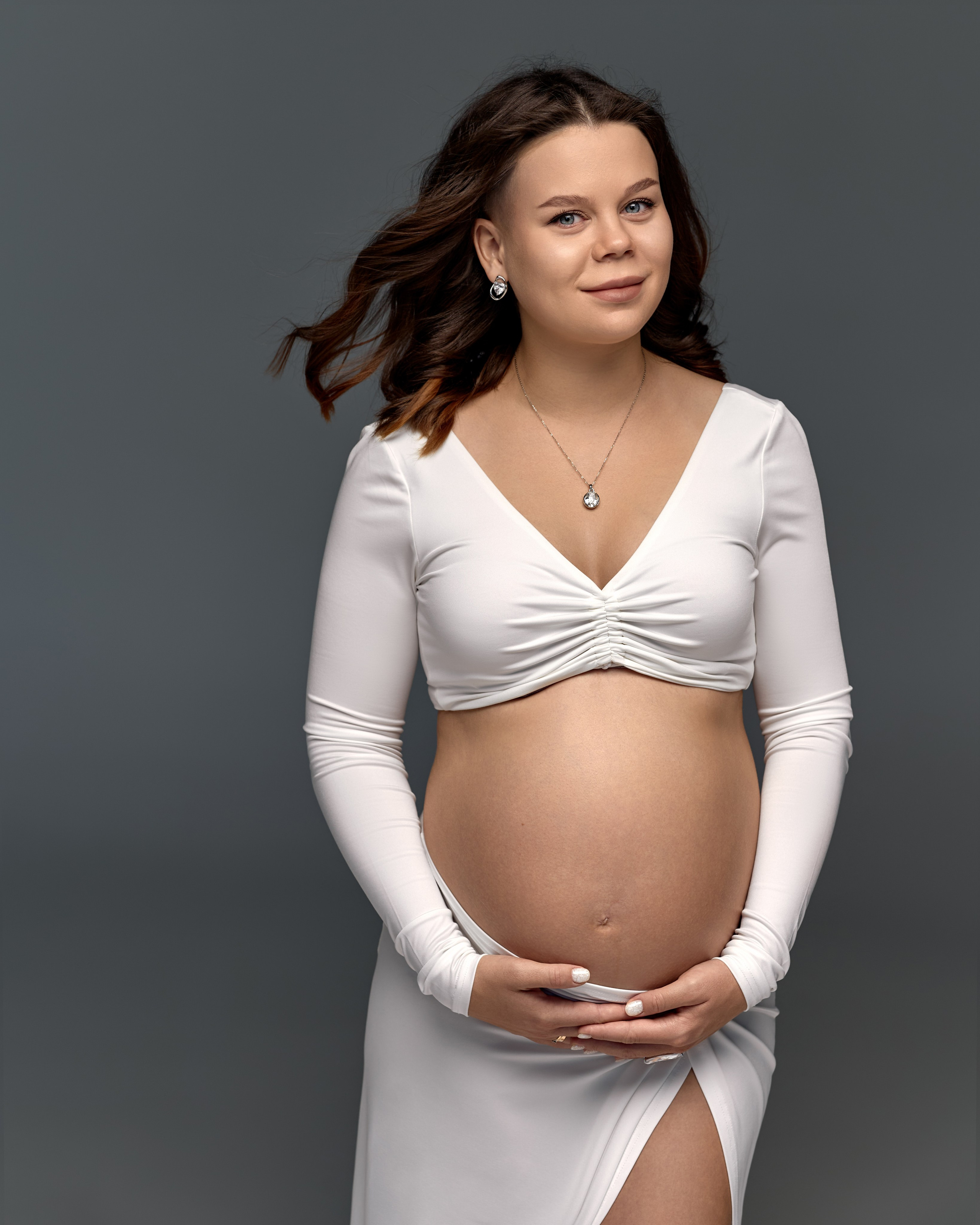 Фотосессия - будущая мама в белом платье с нежной улыбкой, держит живот. Профессиональная студийная съёмка для запоминающихся моментов ожидания ребёнка.