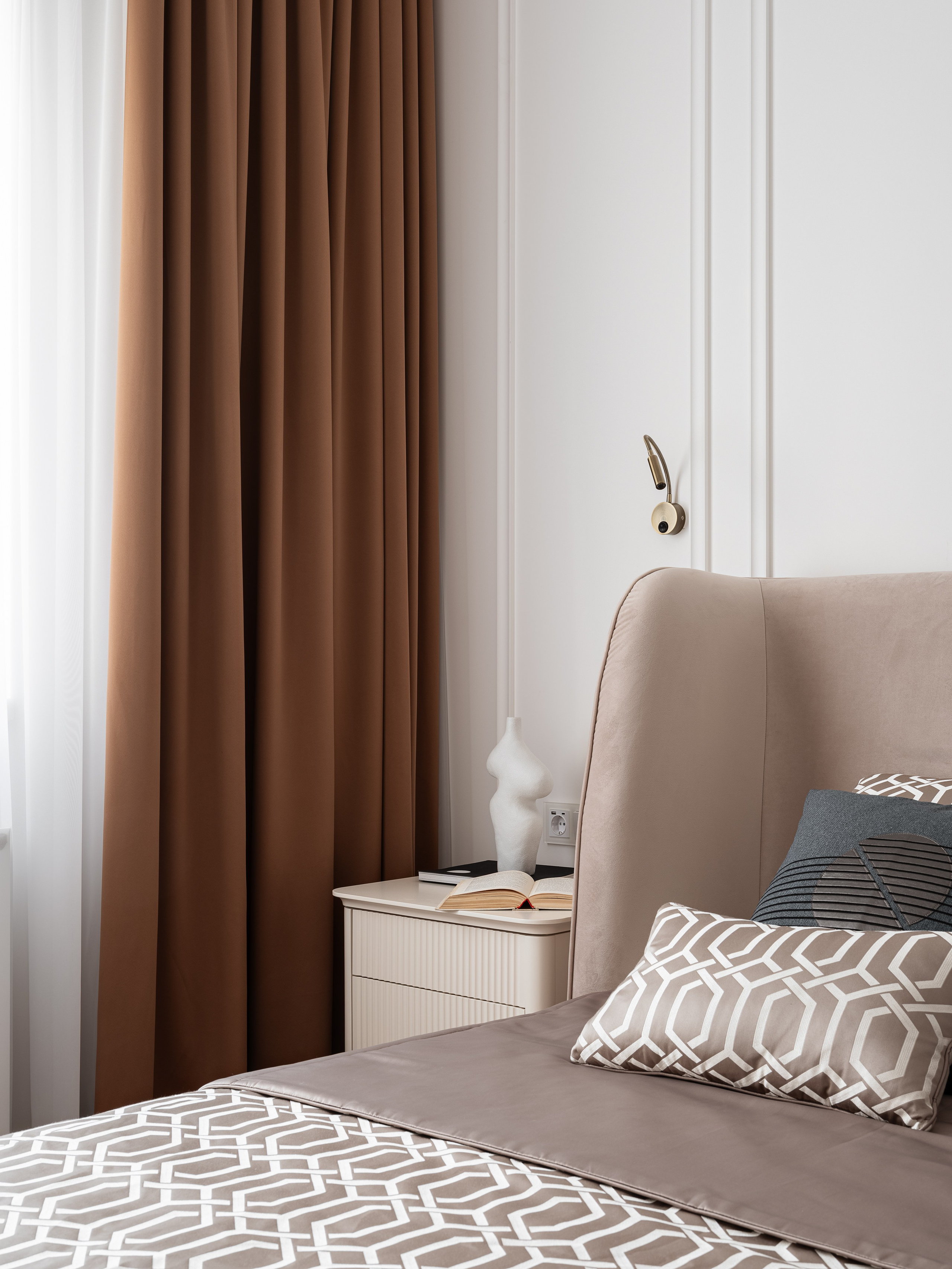 Современная спальня в спокойных тонах с мягким изголовьем кровати, серыми подушками с геометрическим рисунком, прикроватной тумбой и белой вазой на фоне коричневых штор, созданная дизайнером Василиссой Бачевой, стилистом Натальей Якушевой и запечатленная фотографом Сергеем Денисенко.