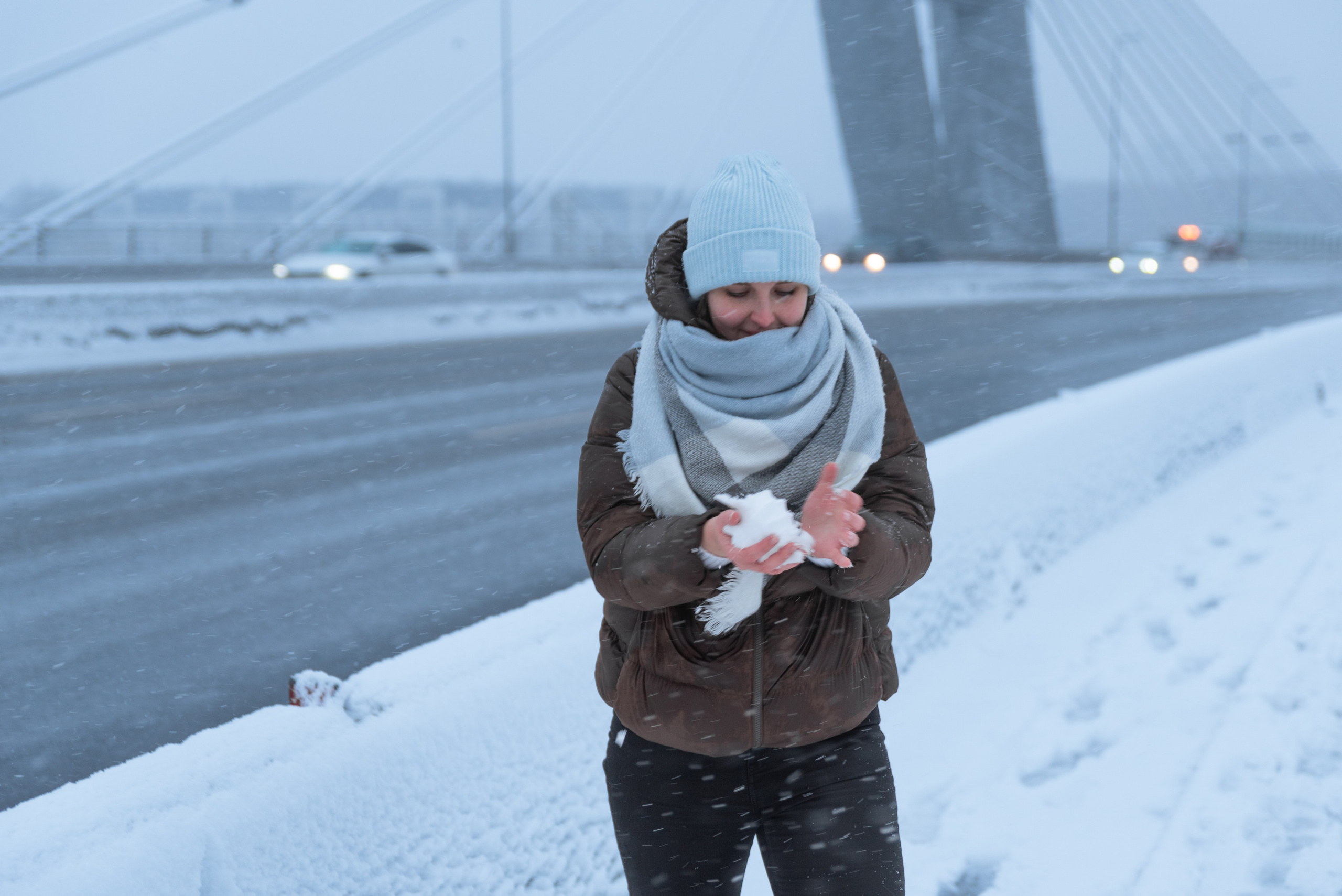 Девушка стоит на мосту, зима, за спиной широкая дорога и опора моста. Девушка лепит снежок.