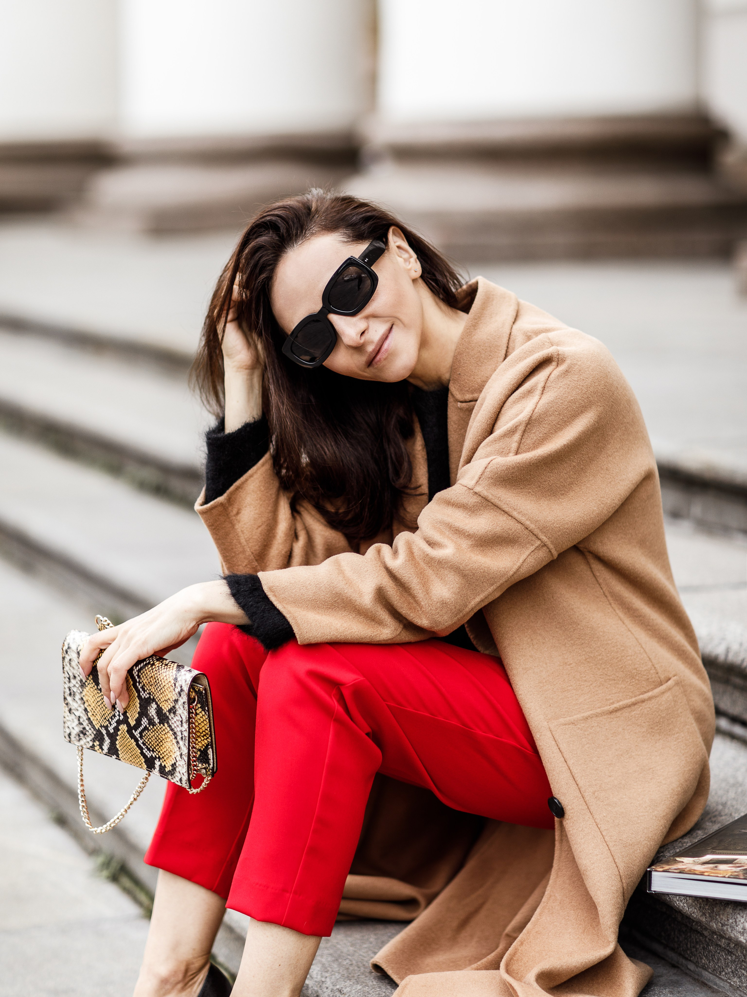 Девушка в солнечных очках, бежевом пальто и красных брюках сидит на ступенях