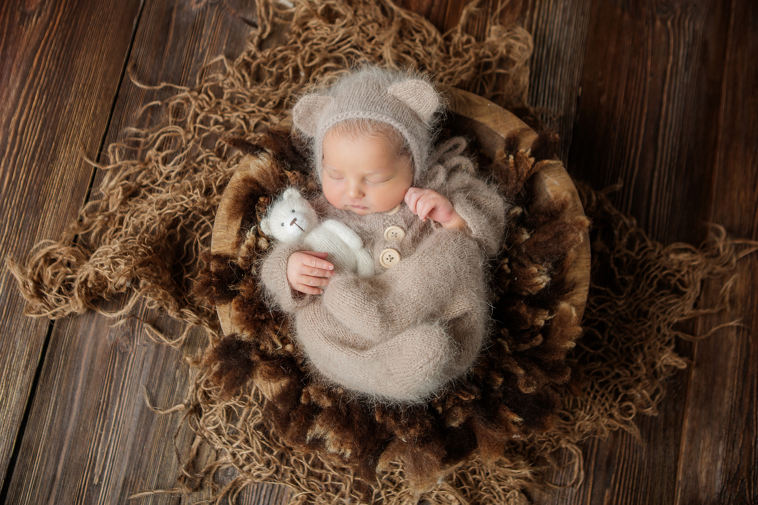 спящий новорожденный малыш в шапочке с ушками с игрушкой котенка в руке в деревянной корзинке на коричневом фоне