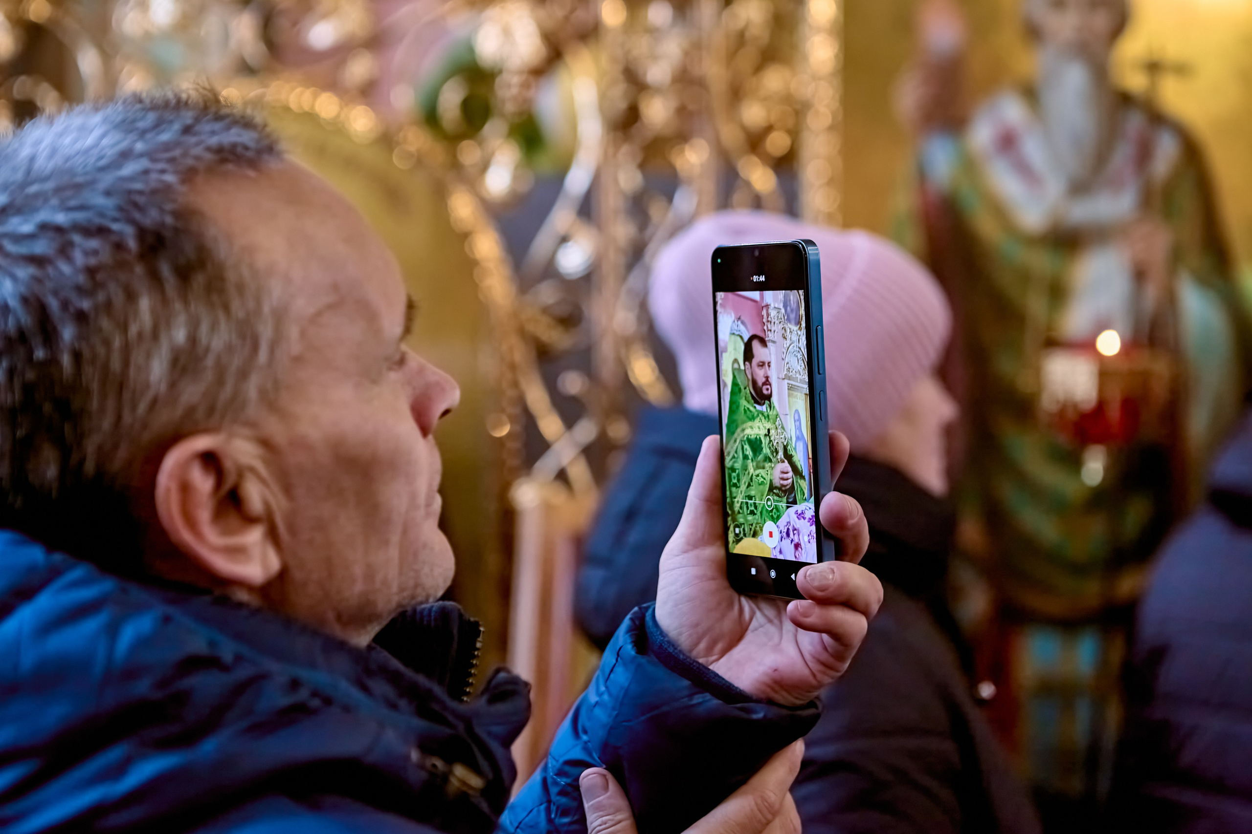 Мужчина с телефоном на котором изображен священник проводящий проповедь