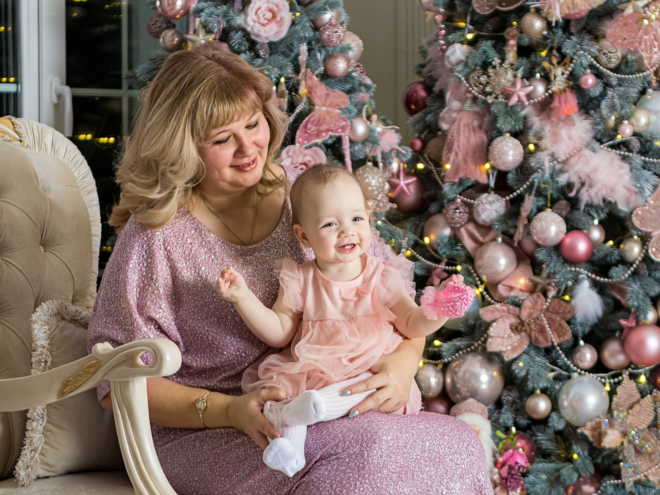 Взрослая женщина сидит в кресле на фоне новогодней елки а на руках у нее внучка радостно улыбается