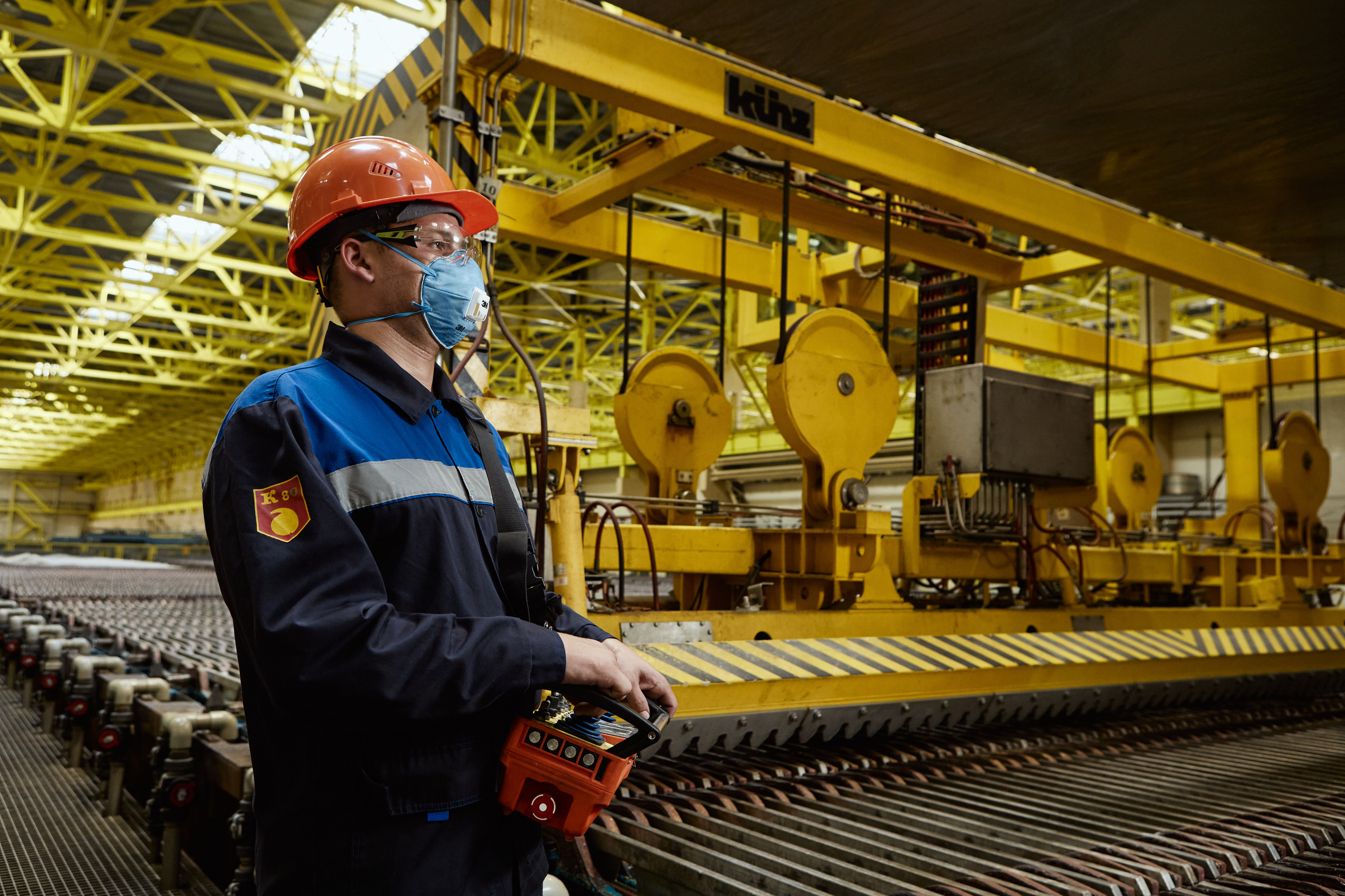 Рабочий в оранжевой каске и респираторе на фоне электролизного цеха