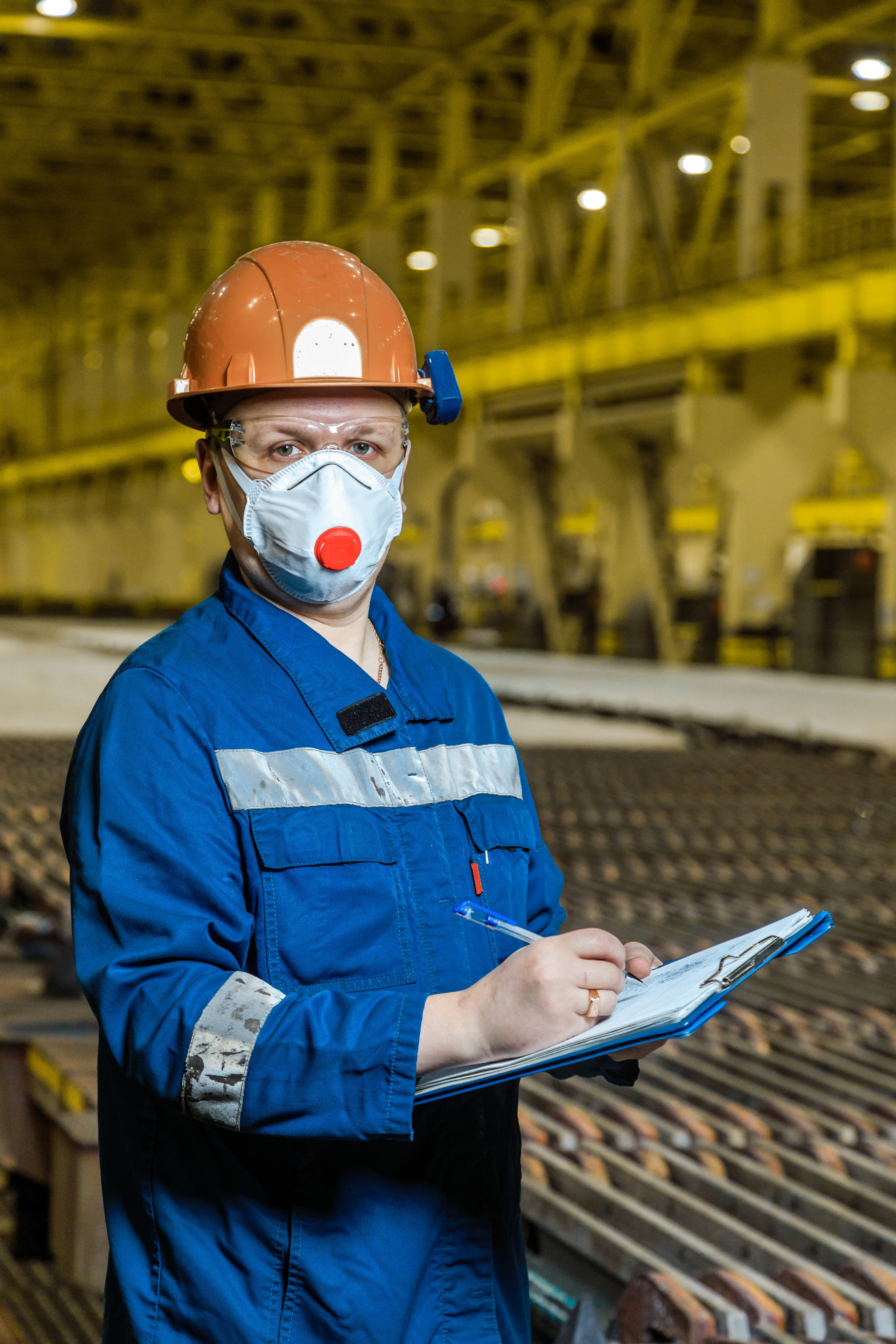 Сотрудник предприятия в каске, спецодежде и респираторе держит в руках бумаги
