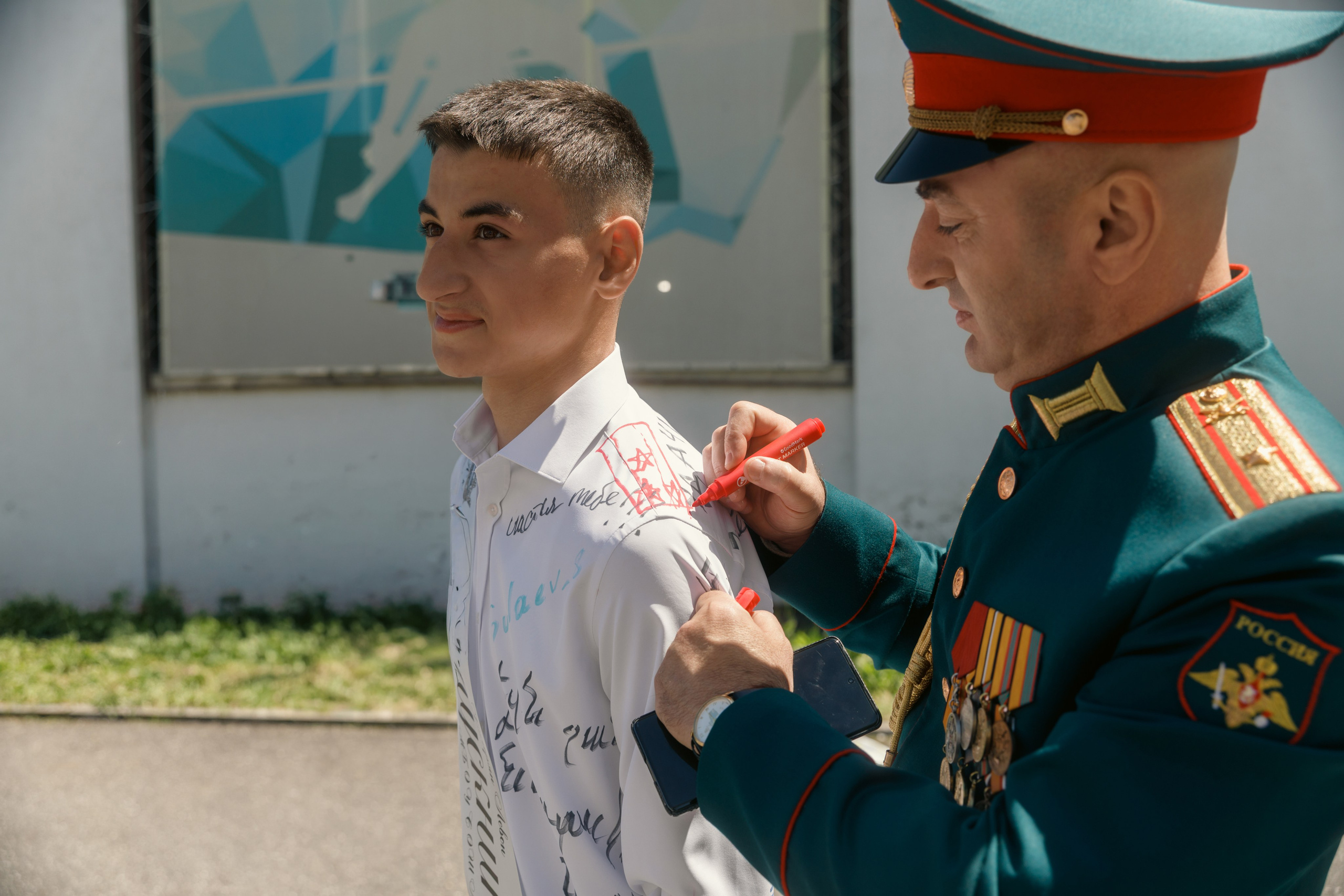 Военный в парадной форме оставляет пожелания и подпись на рубашке выпускника во время празднования последнего звонка на школьном мероприятии.