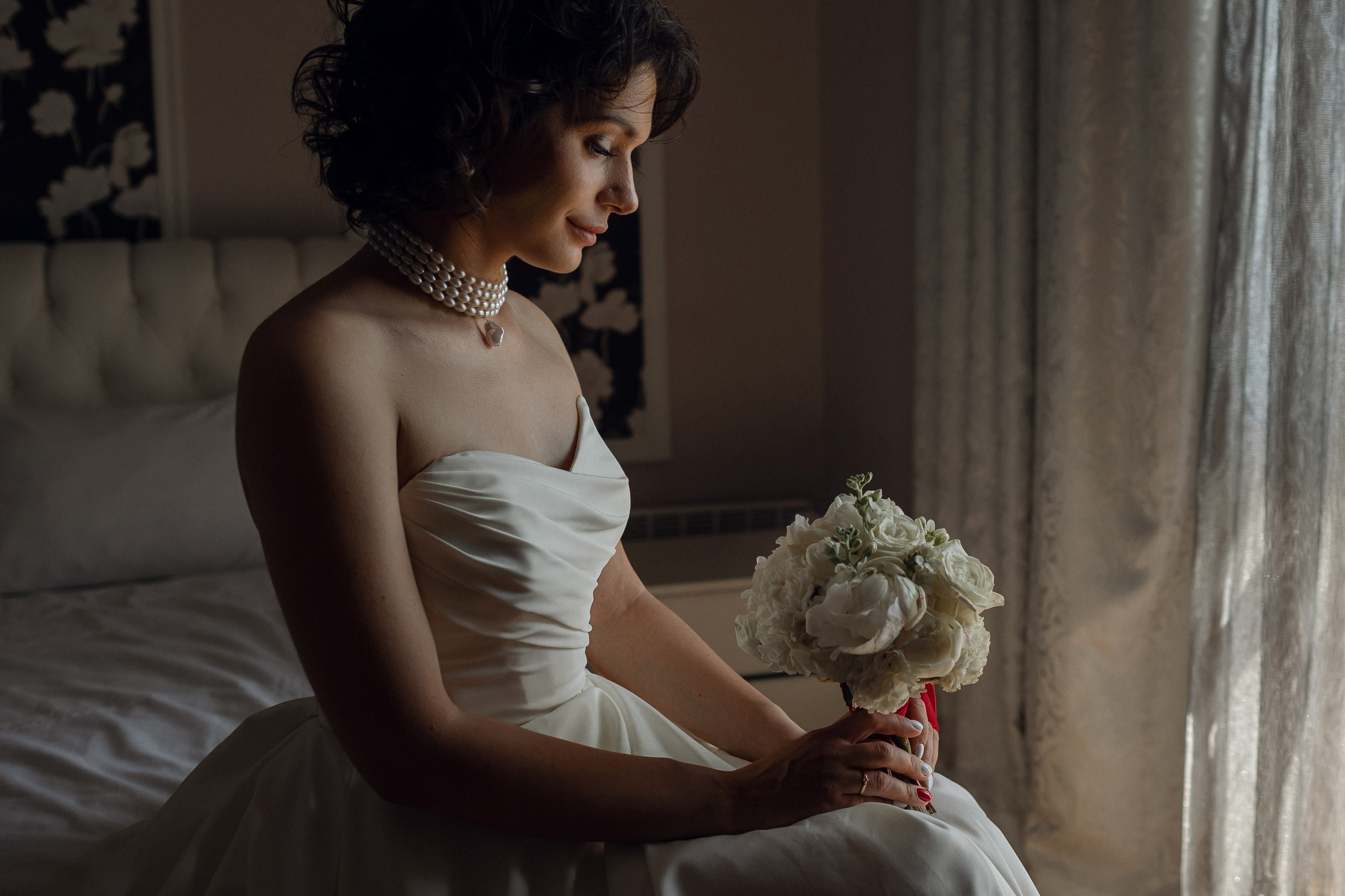 Элегантный свадебный портрет в полумраке: невеста в атласном платье сидит на кровати , держа букет роз при мягком свете от окна