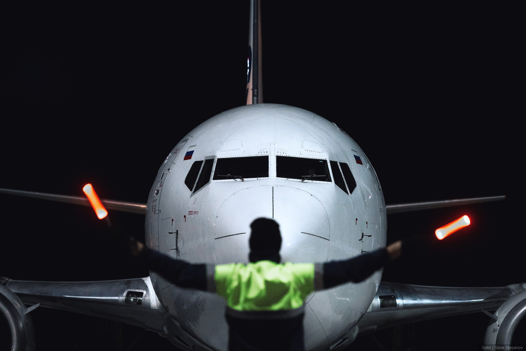 Носовая часть пассажирского лайнера Boeing 737 на перроне аэродрома в темное время суток. На переднем плане — авиационный маршал со спины с разведенными в стороны светящимися сигнальными жезлами. Снимок Gelio.