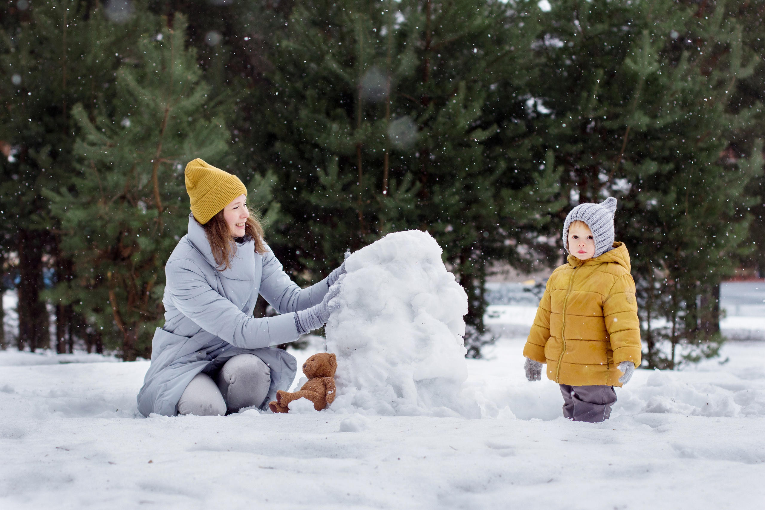 Мама и дочка лепят снеговика с в снежном сосновом лесу