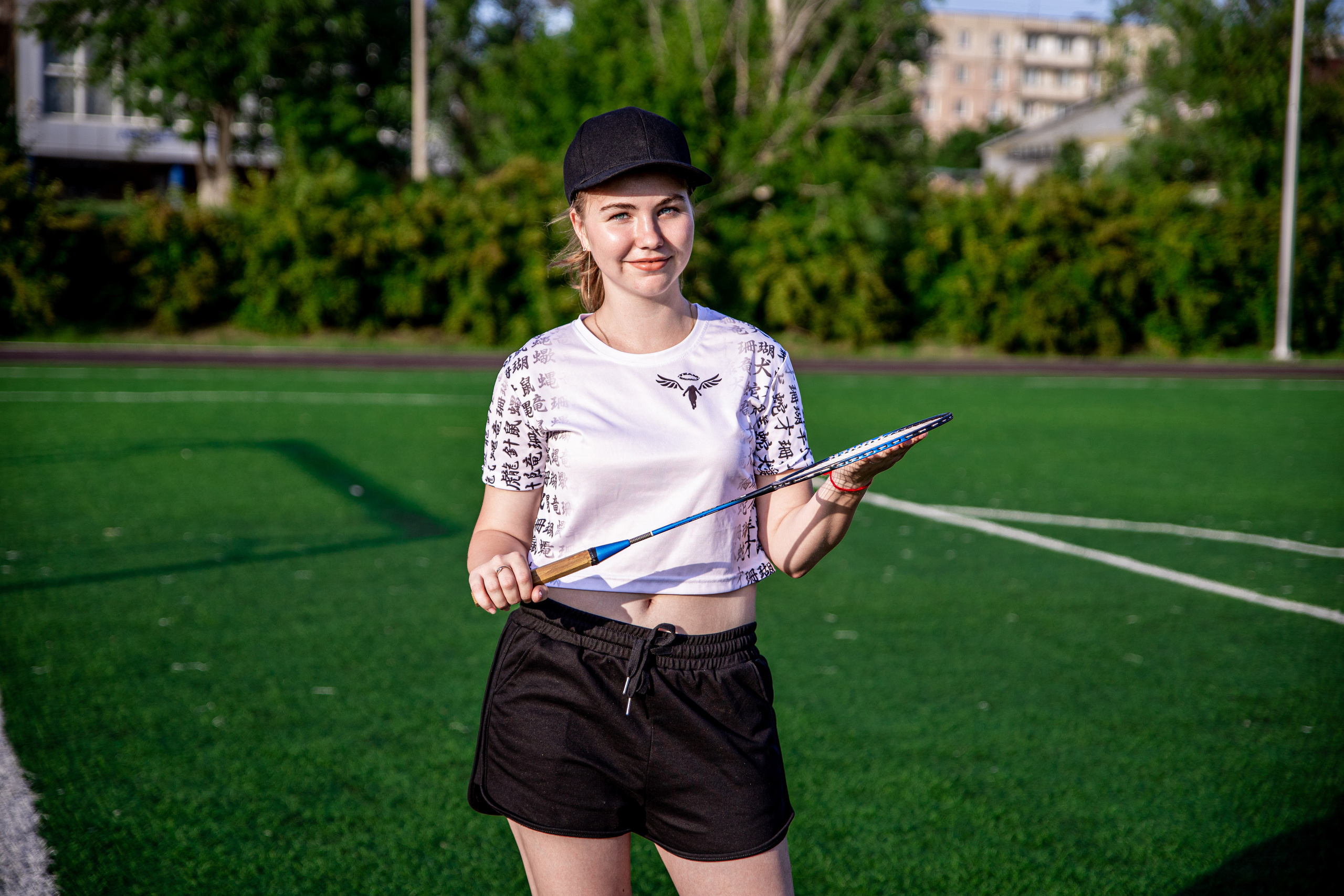 Девушка стоит на стадионе на зелёном фоне, в кепке, в  белой футболке, с ракеткой в руке