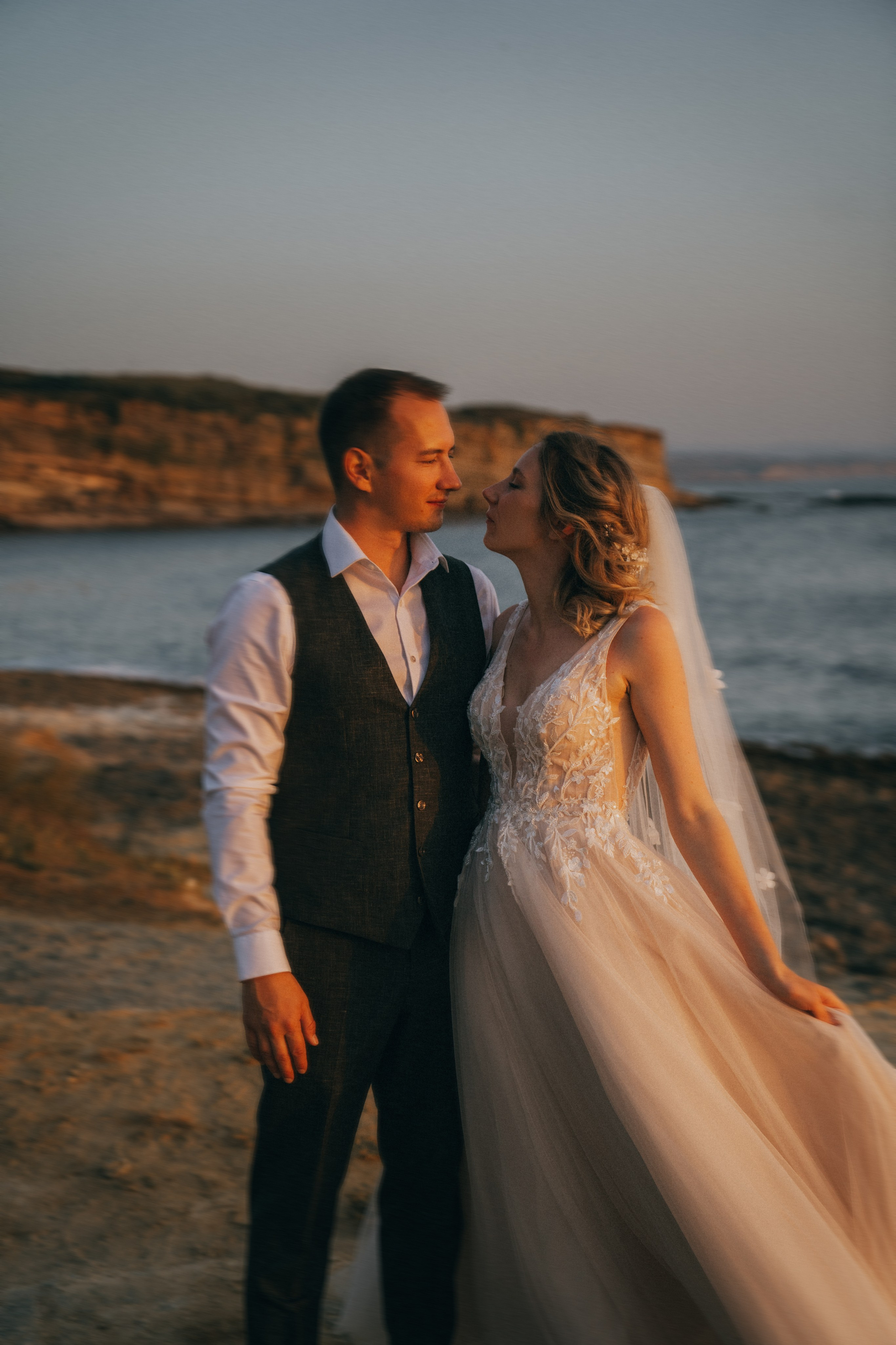 Счастливая пара жених и невеста целуется на фоне волн  скал и заката у океана.