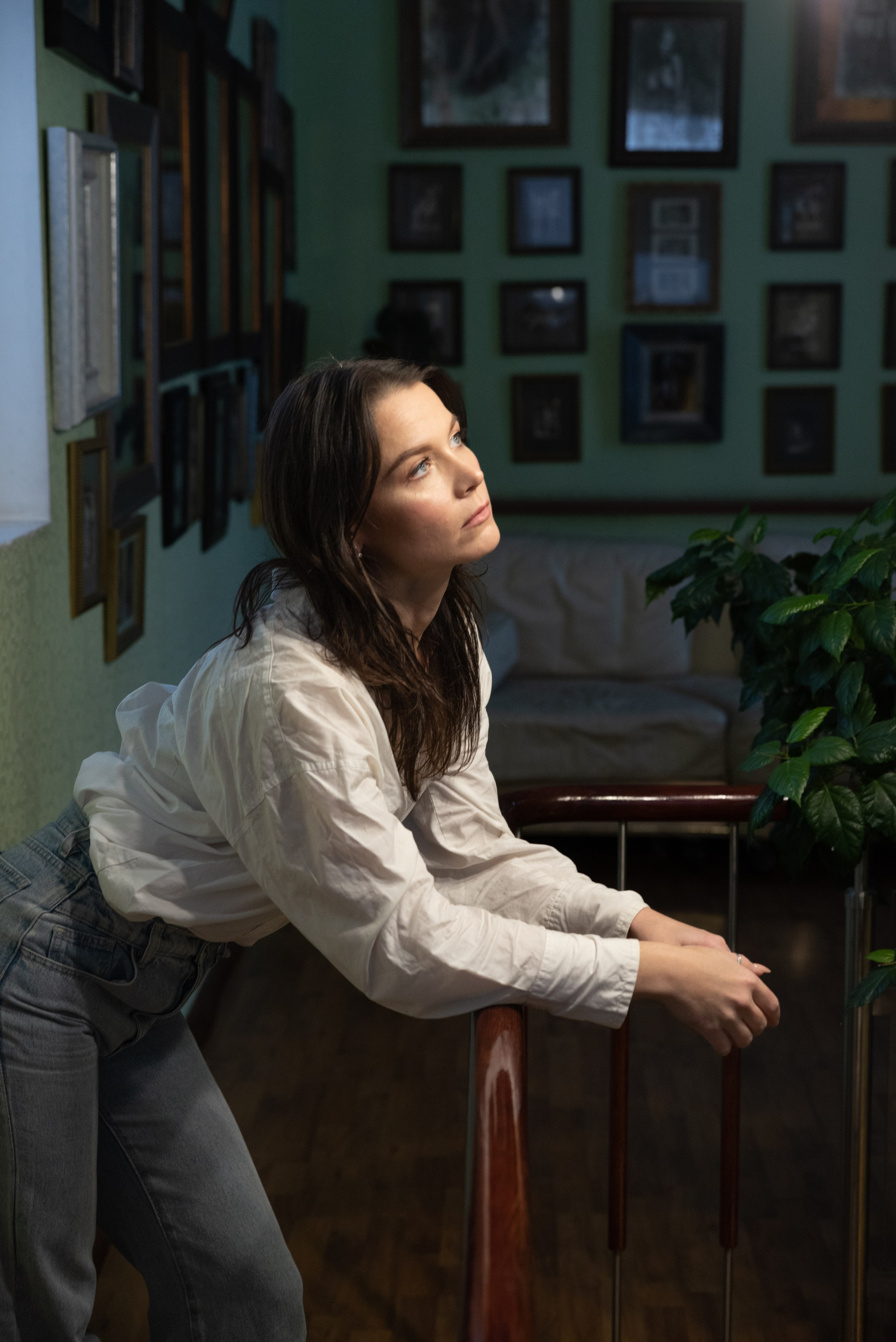 Комната, на стенах множество фотографий в рамках, на переднем плане молодая женщина, стоит облокотившись на перила лестницы. Сверху на лицо падает свет.