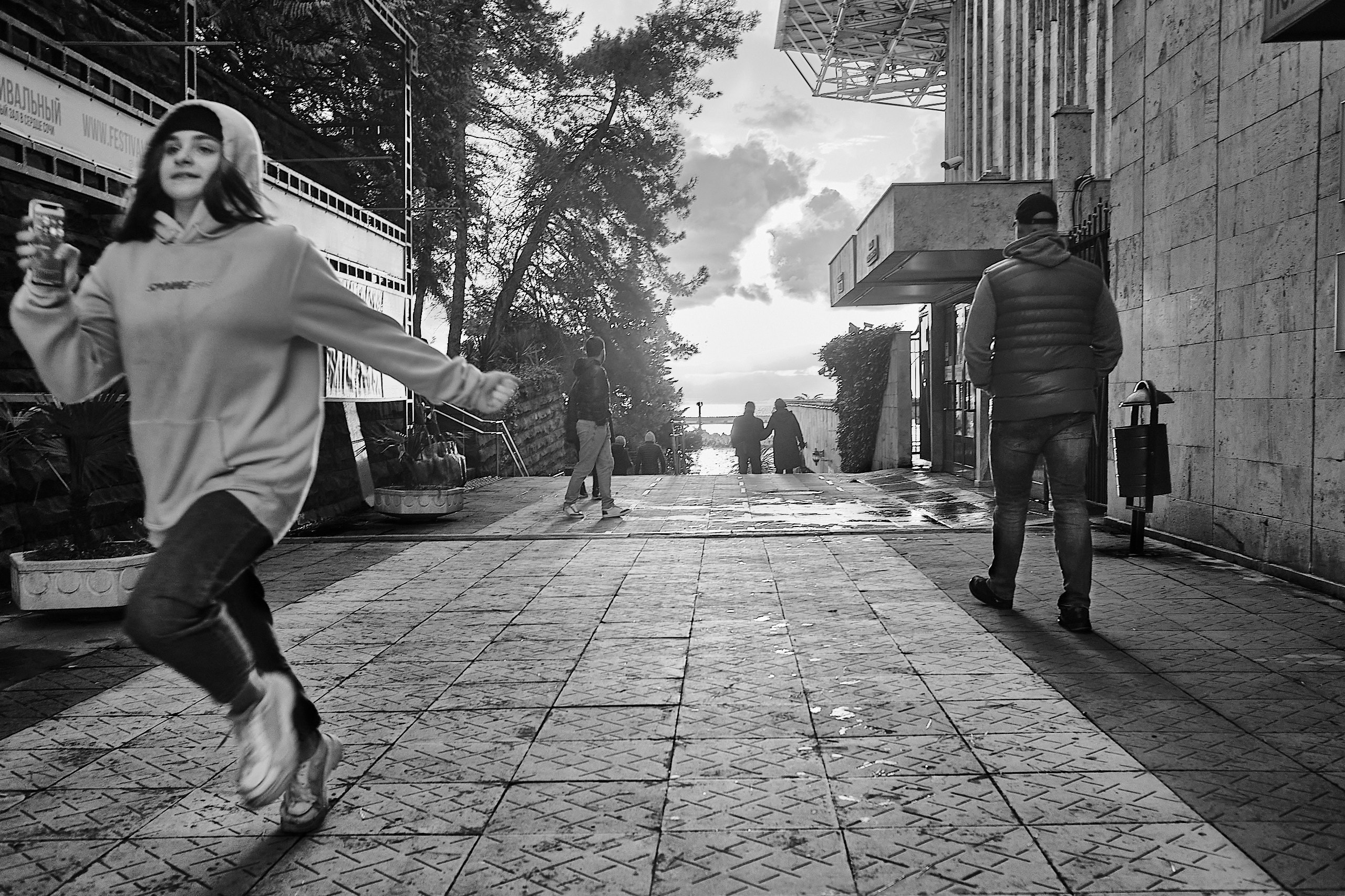 Street view in Sochi, near the Festivalny Concert Hall. The setting sun shines through the clouds in a street perspective. The sea is visible. In the foreground, a girl runs out of the frame. Passers-by are visible in the distance. (Вид на улицу в Сочи, возле концертного зала "Фестивальный". Лучи заходящего солнца пробиваются сквозь облака в перспективе улицы. Виднеется море. На переднем плане девушка выбегает из кадра. В далеке видны прохожие.)