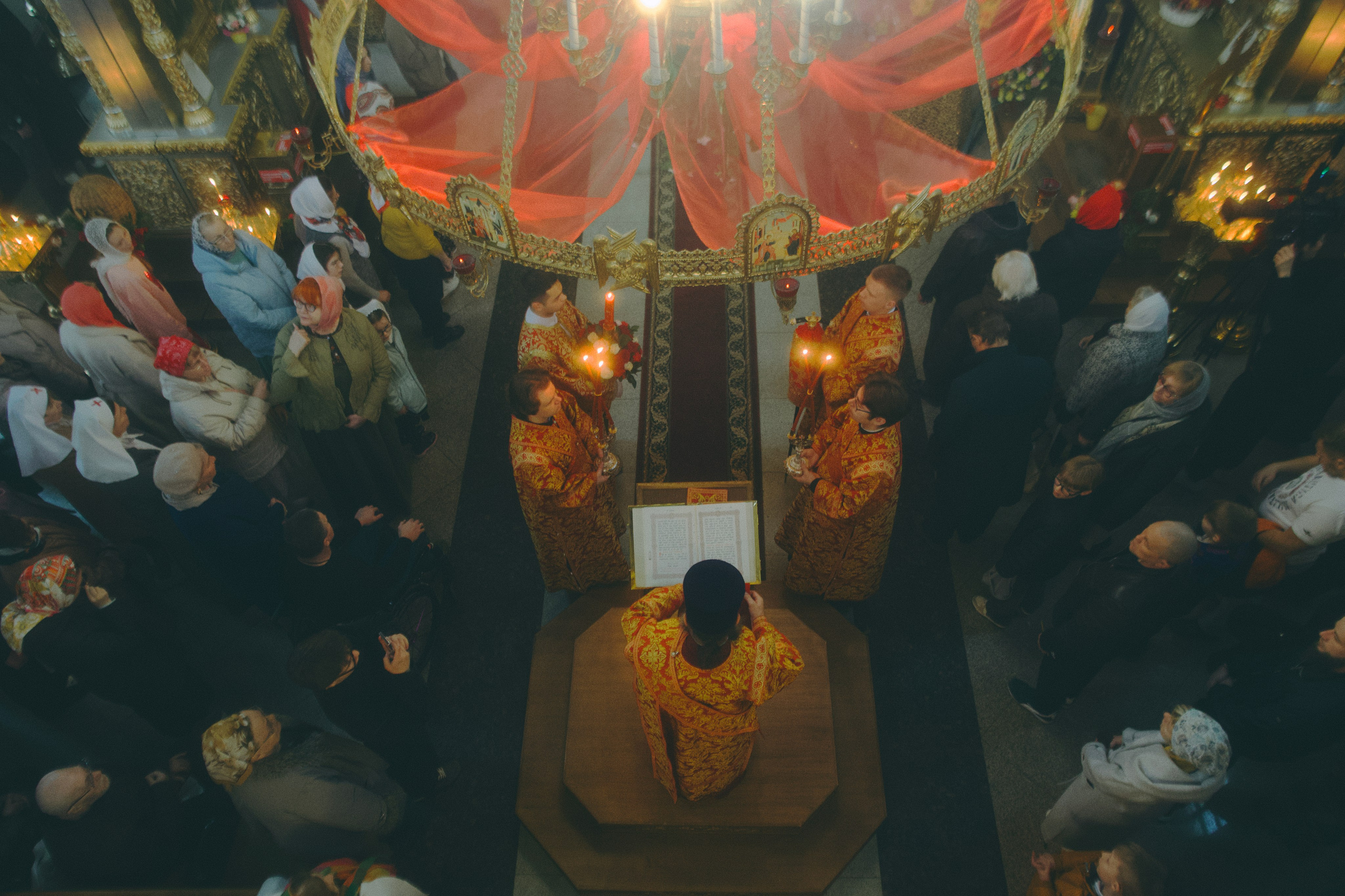 Вид сверху на пасхальное богослужение: священники в золотых облачениях вокруг аналоя, прихожане молятся под большим паникадилом с красным убранство