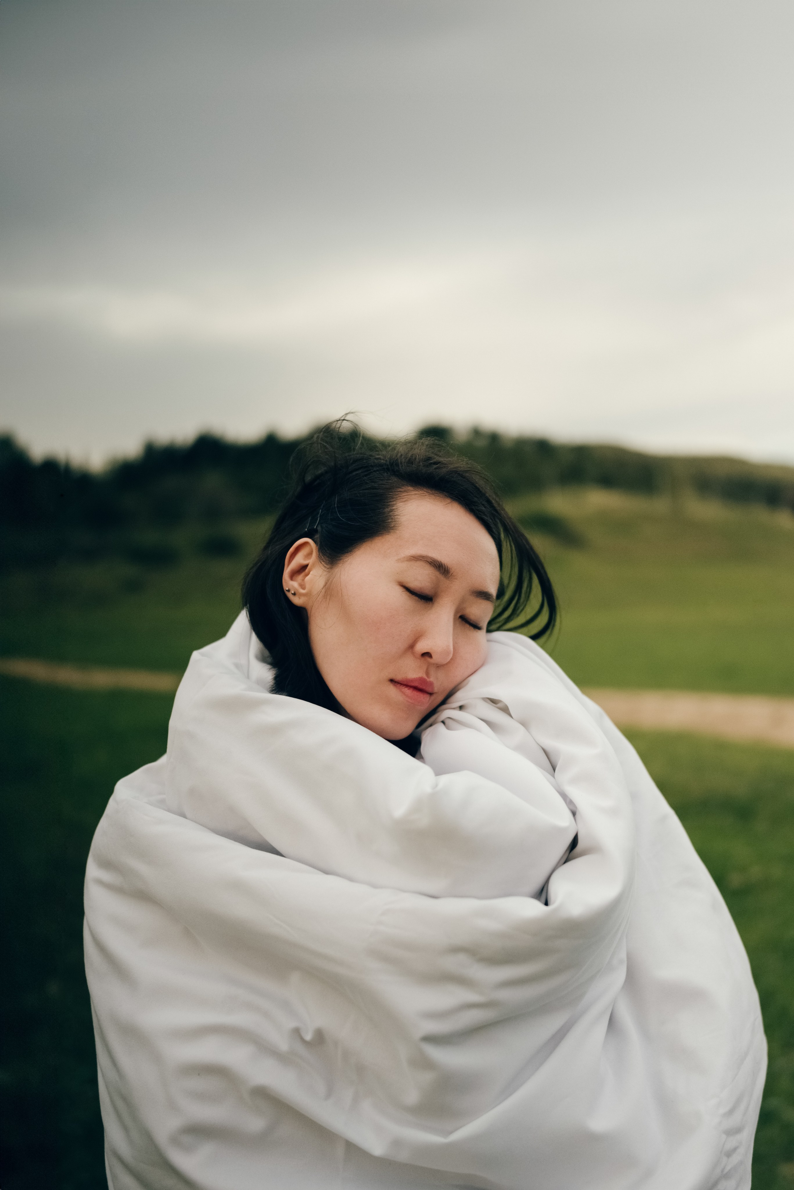 Портрет азиатской девушки в одеяле на фоне зеленого поля. Модель расслаблена и укутана в теплое белое одеяло