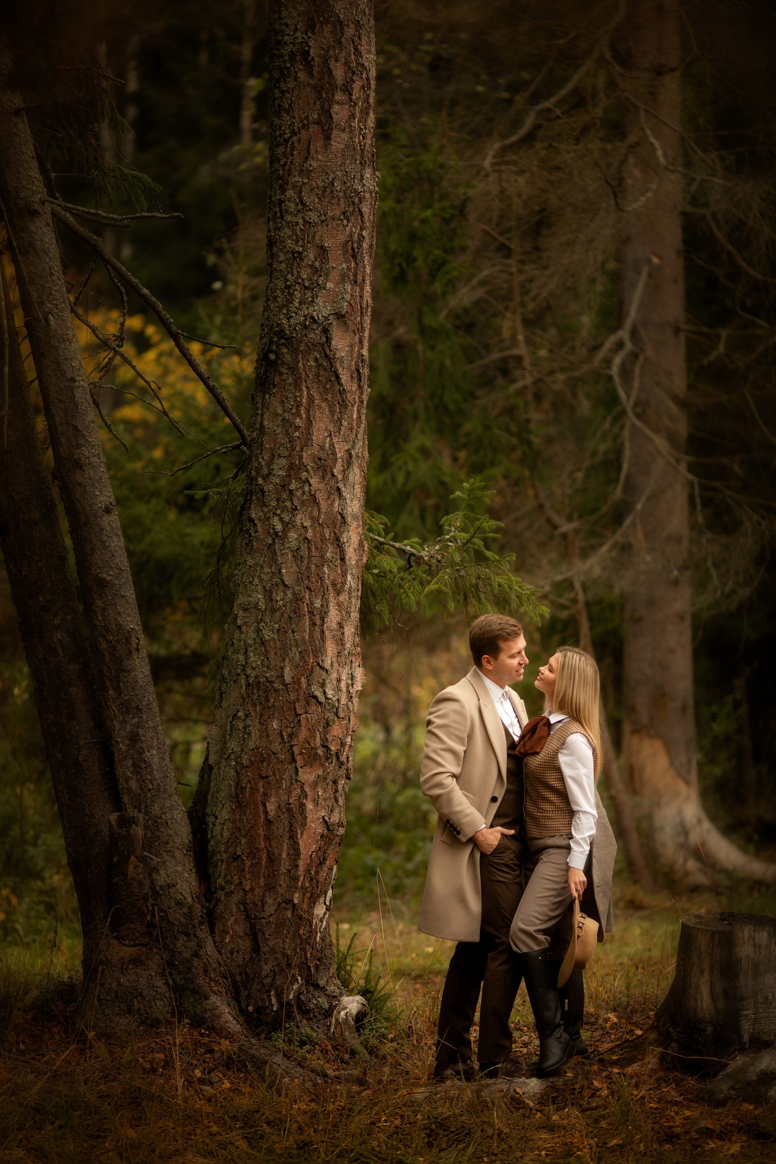 Пара стоит у дерева в осеннем лесу, смотрят друг на друга с теплом, в мягком рассеянном свете среди тёплых коричневых оттенков природы.