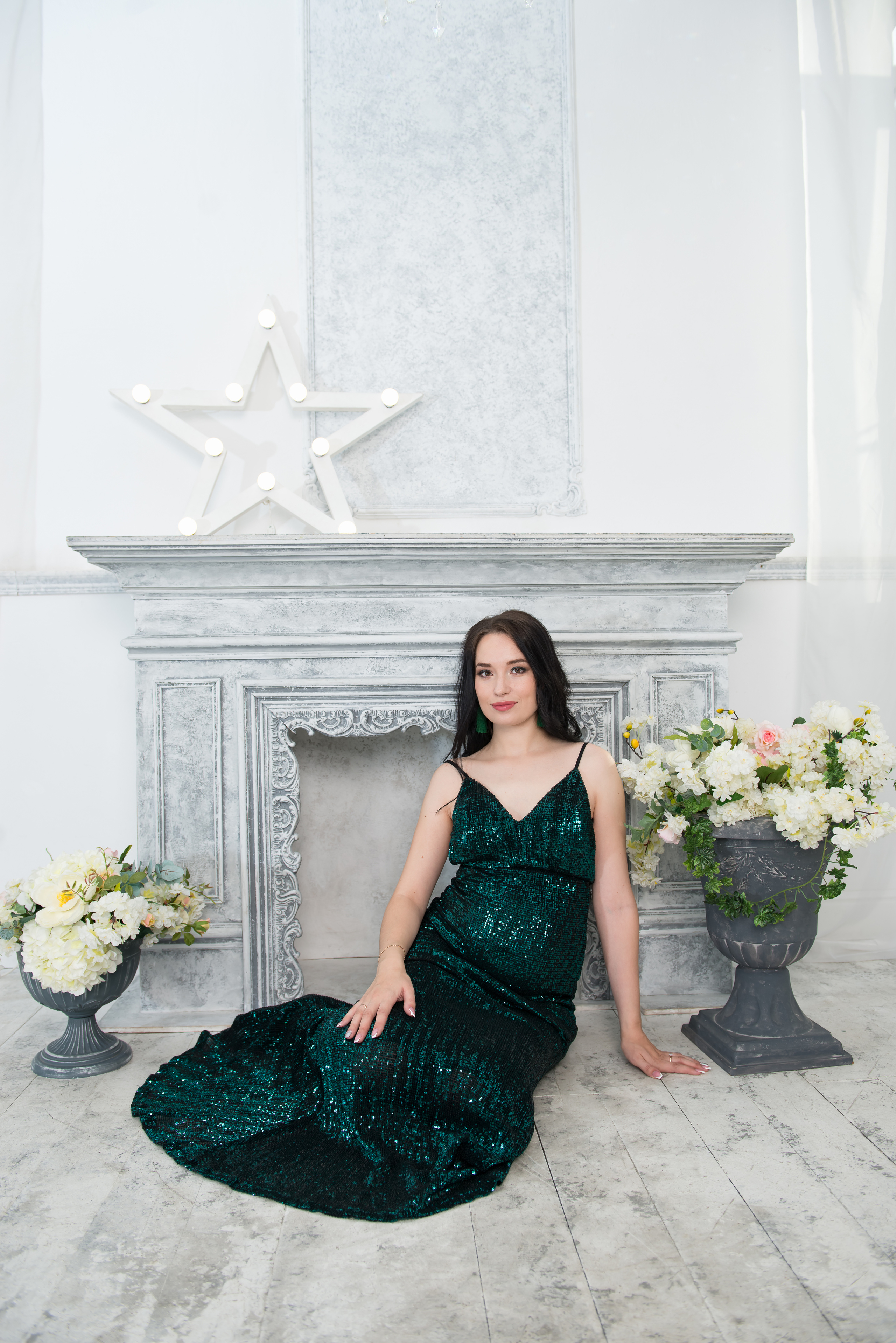 Беременная женщина сидит на полу в фотостудии в зеленом силуэтном платье на фоне камина