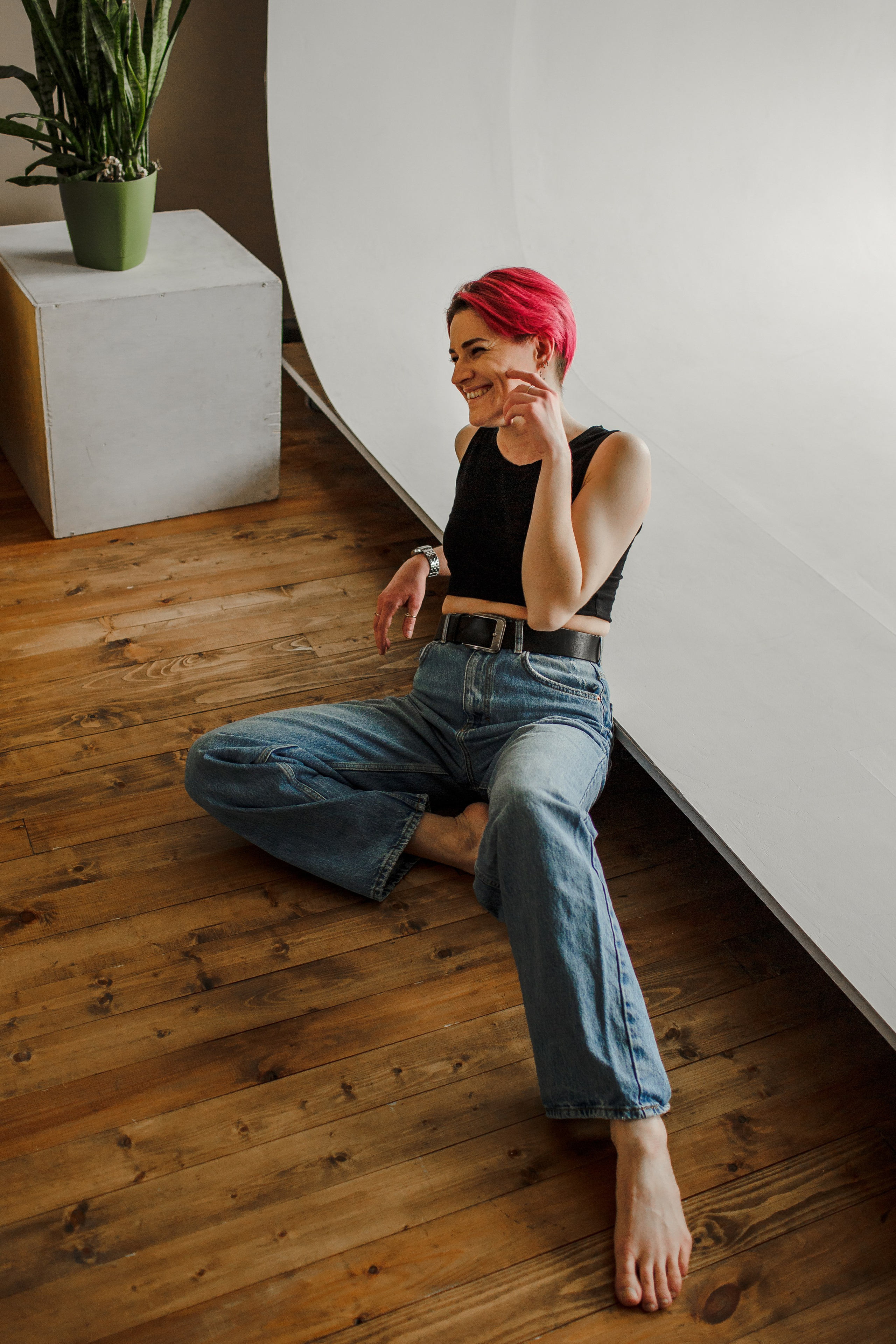 Модель сидит на деревянном полу с белой циклорамой; она вытянула ногу вперед и прикрыла лицо ладонью.