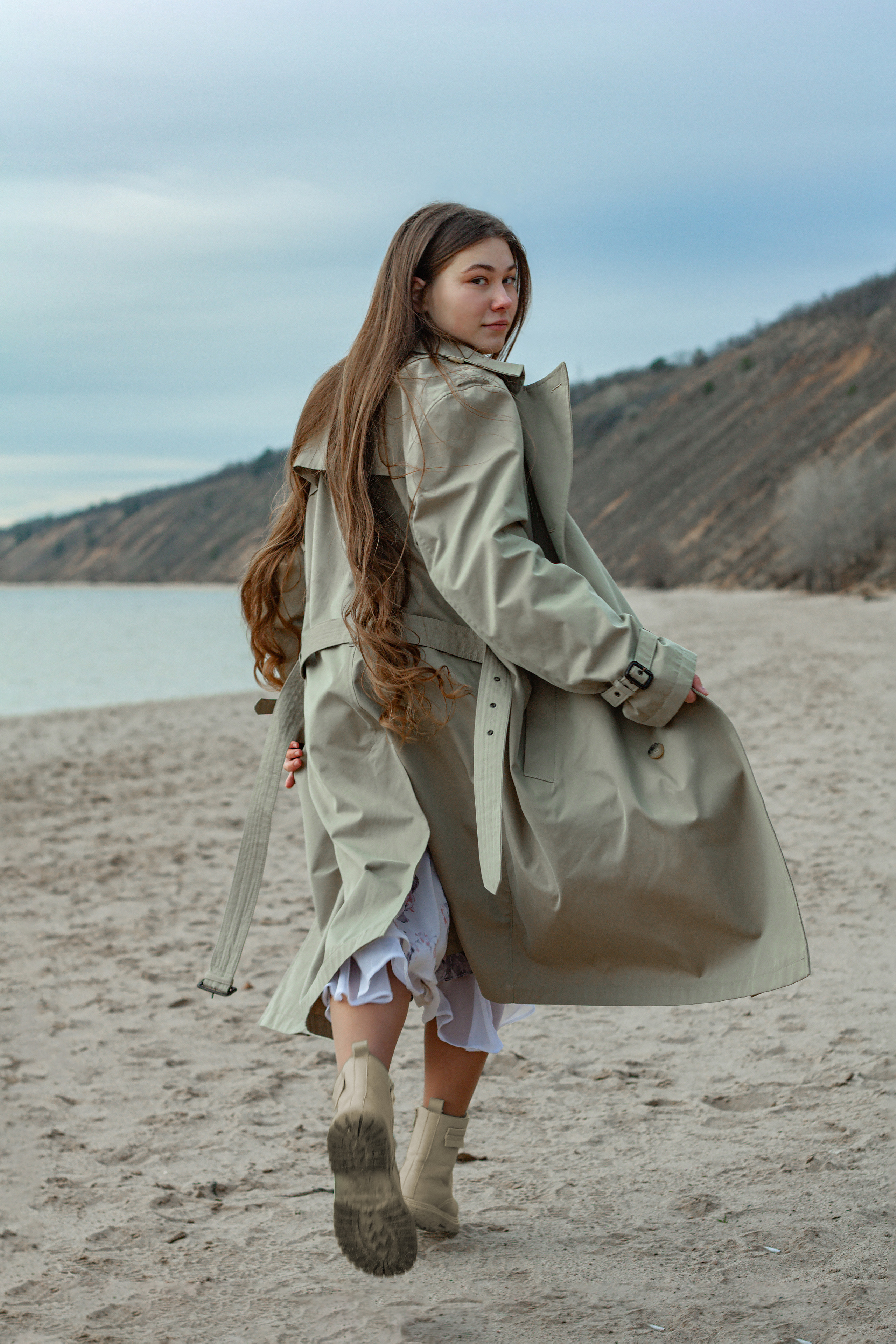 Девушка бежит по песчаному пляжу в светлом пальто.