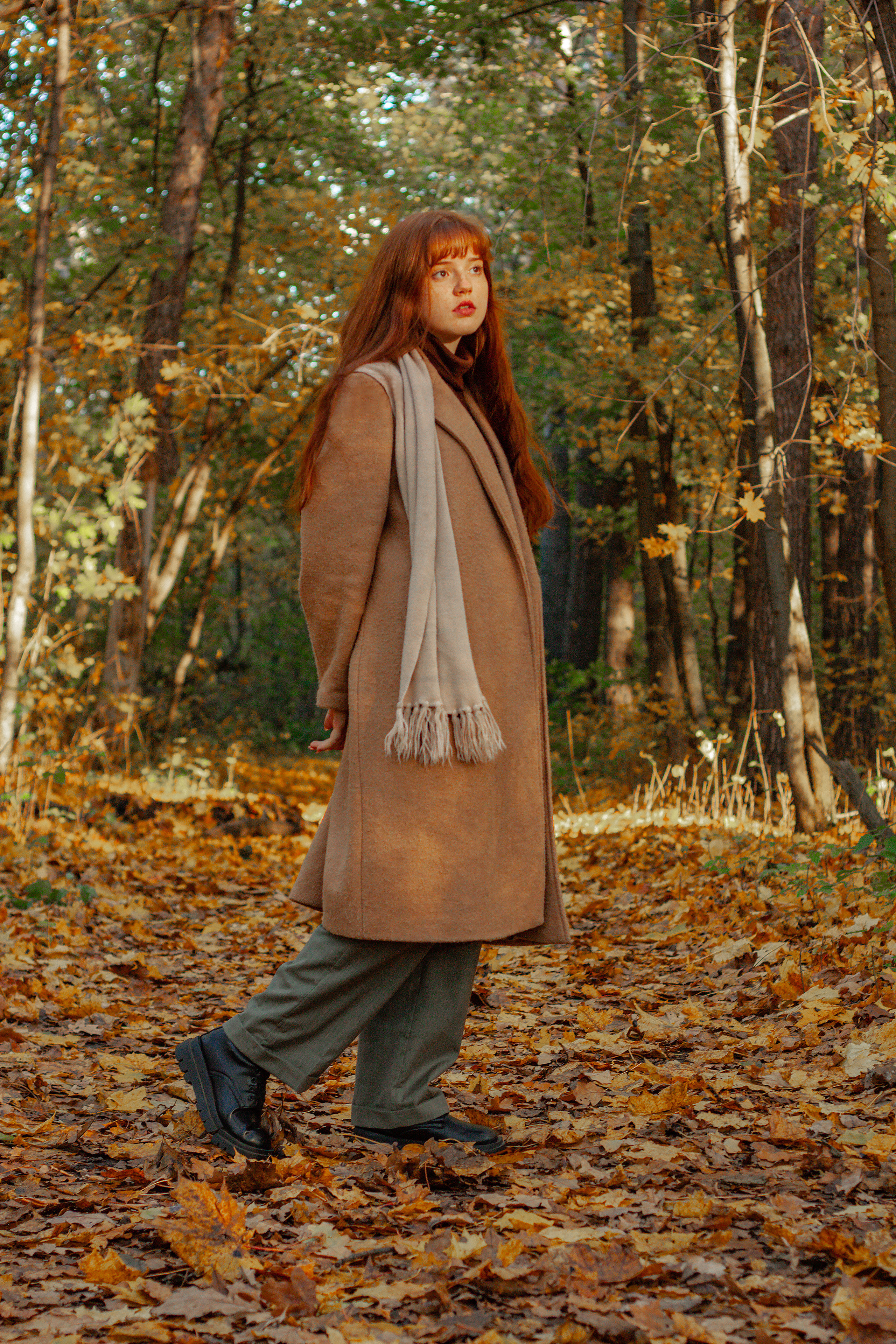 Девушка стоит посреди осеннего леса в пальто и смотрит на солнце. 