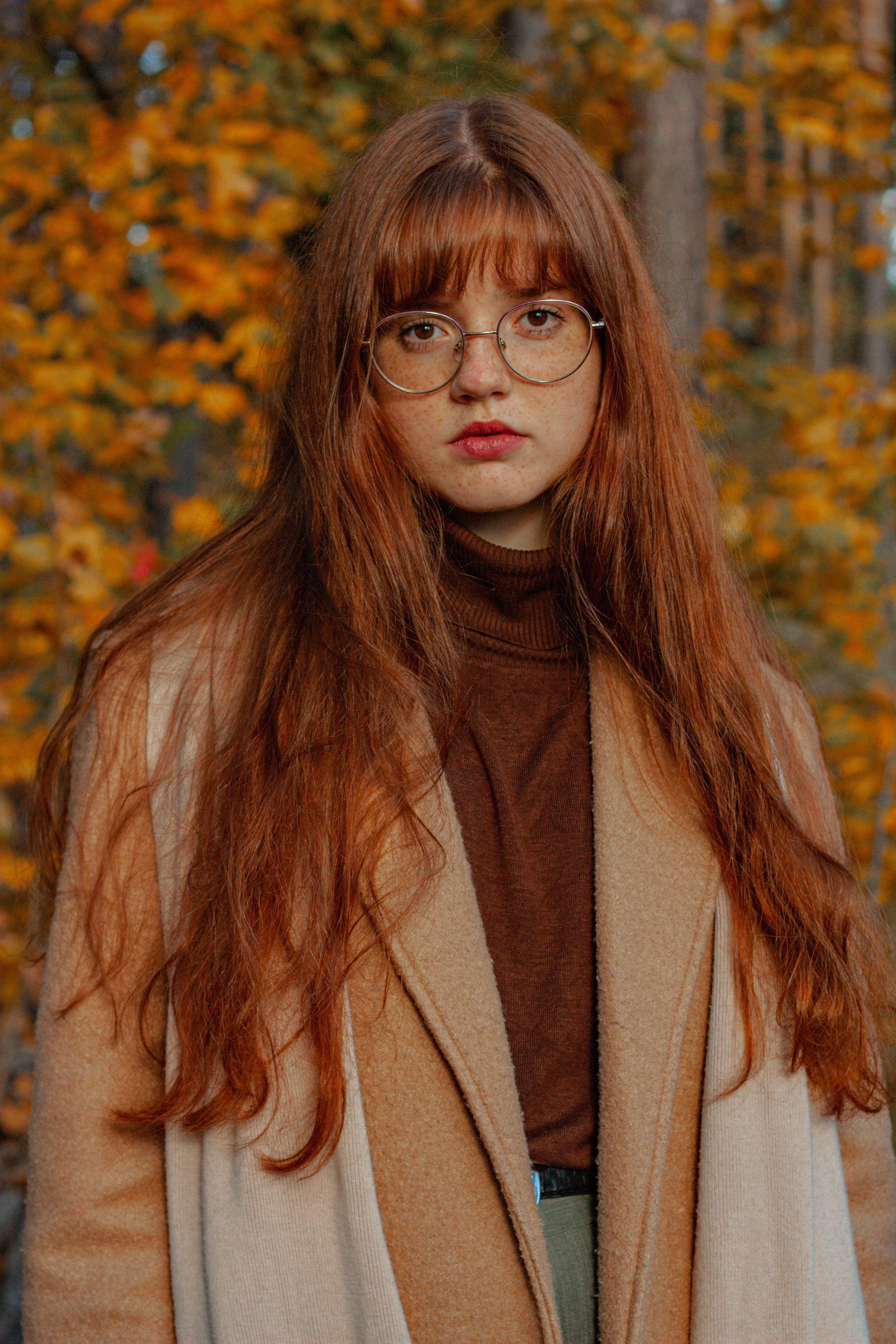 Рыжеволосая девушка в очках в пальто на фоне осеннего леса. 