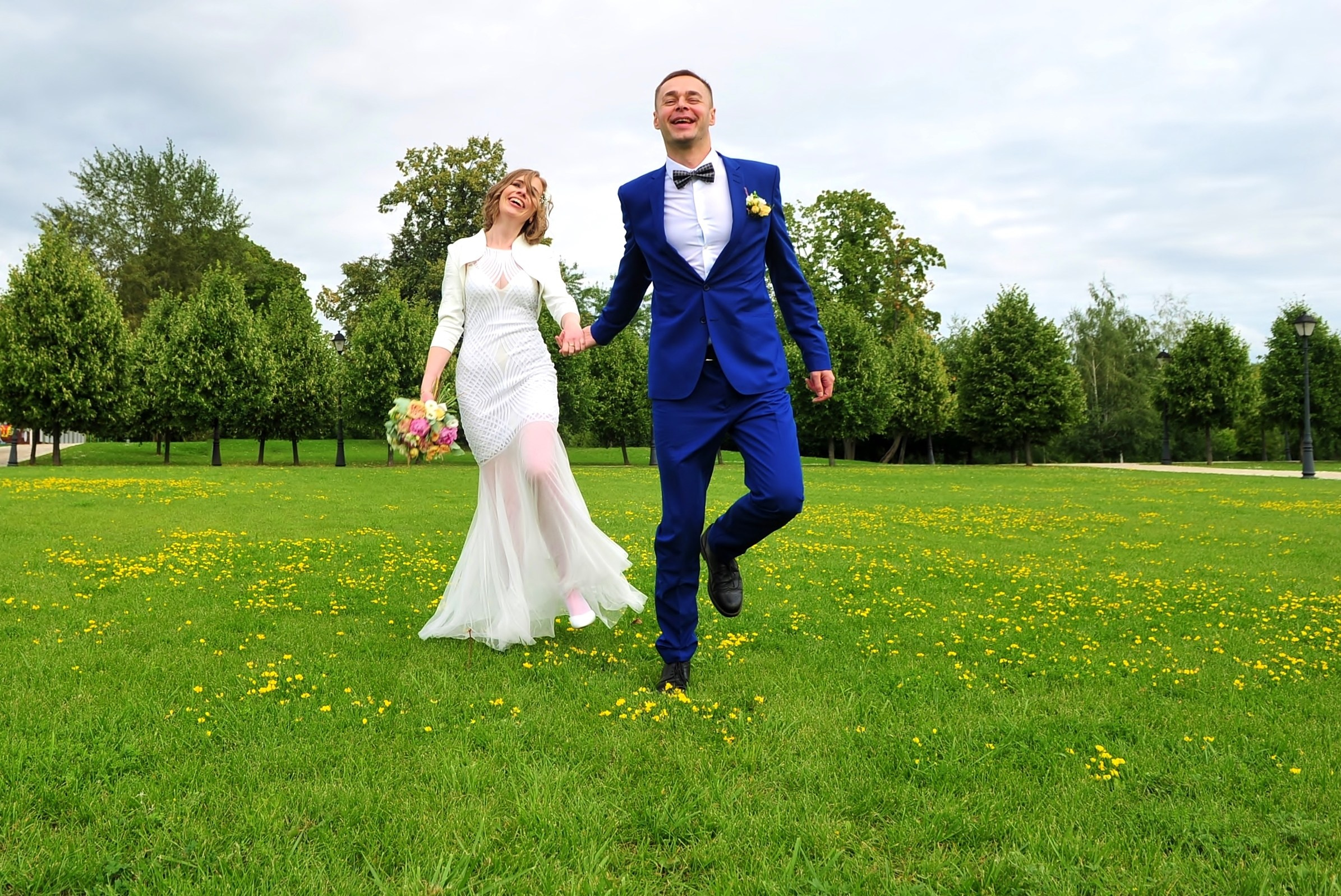 Свадьба в Царицыно хмурым прохладным летним днём, жених ведёт за собой за руку невесту по зеленой лужайке с желтыми одуванчиками