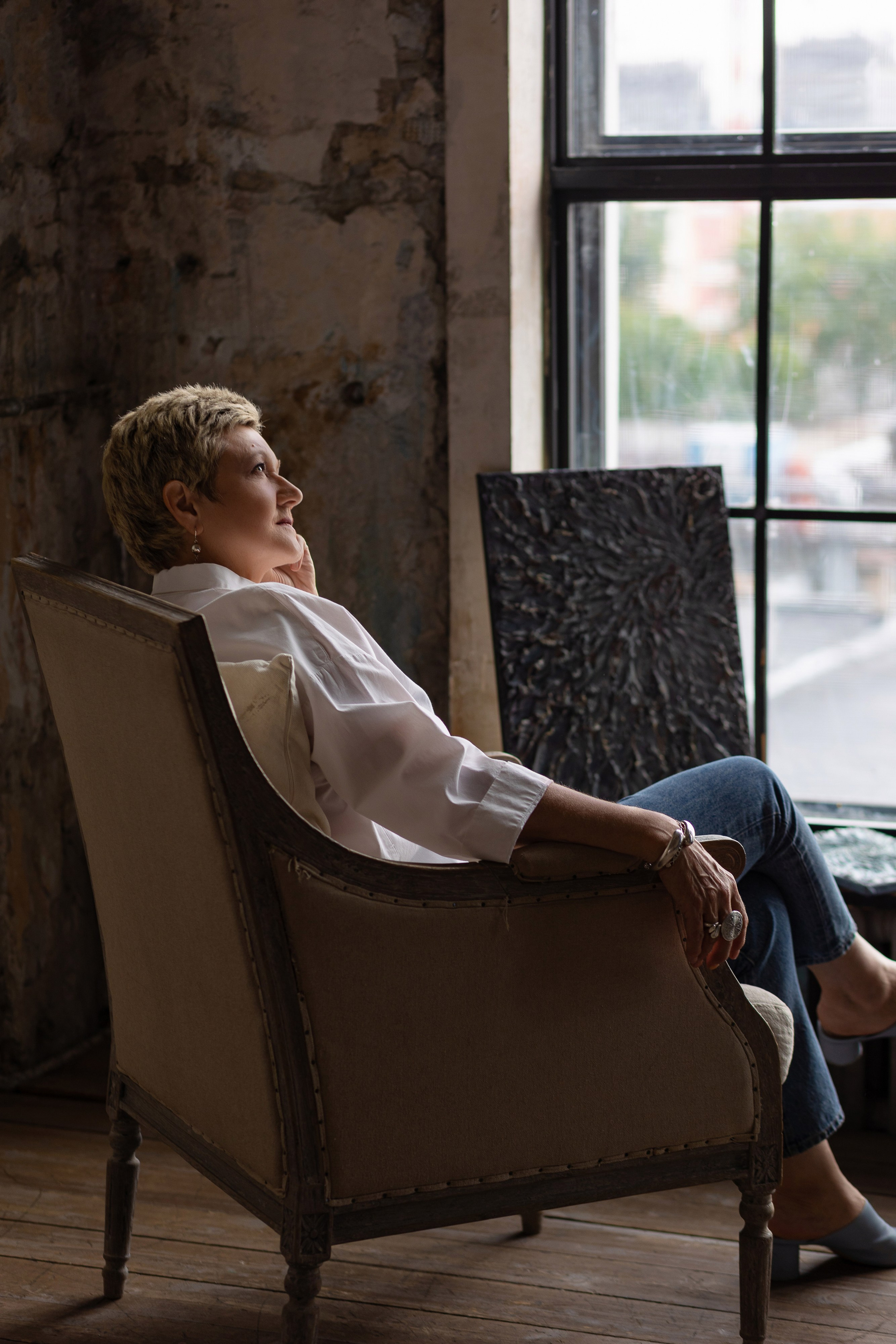 У окна сидит женщина в кресле, правая рука лежит на подлокотнике.