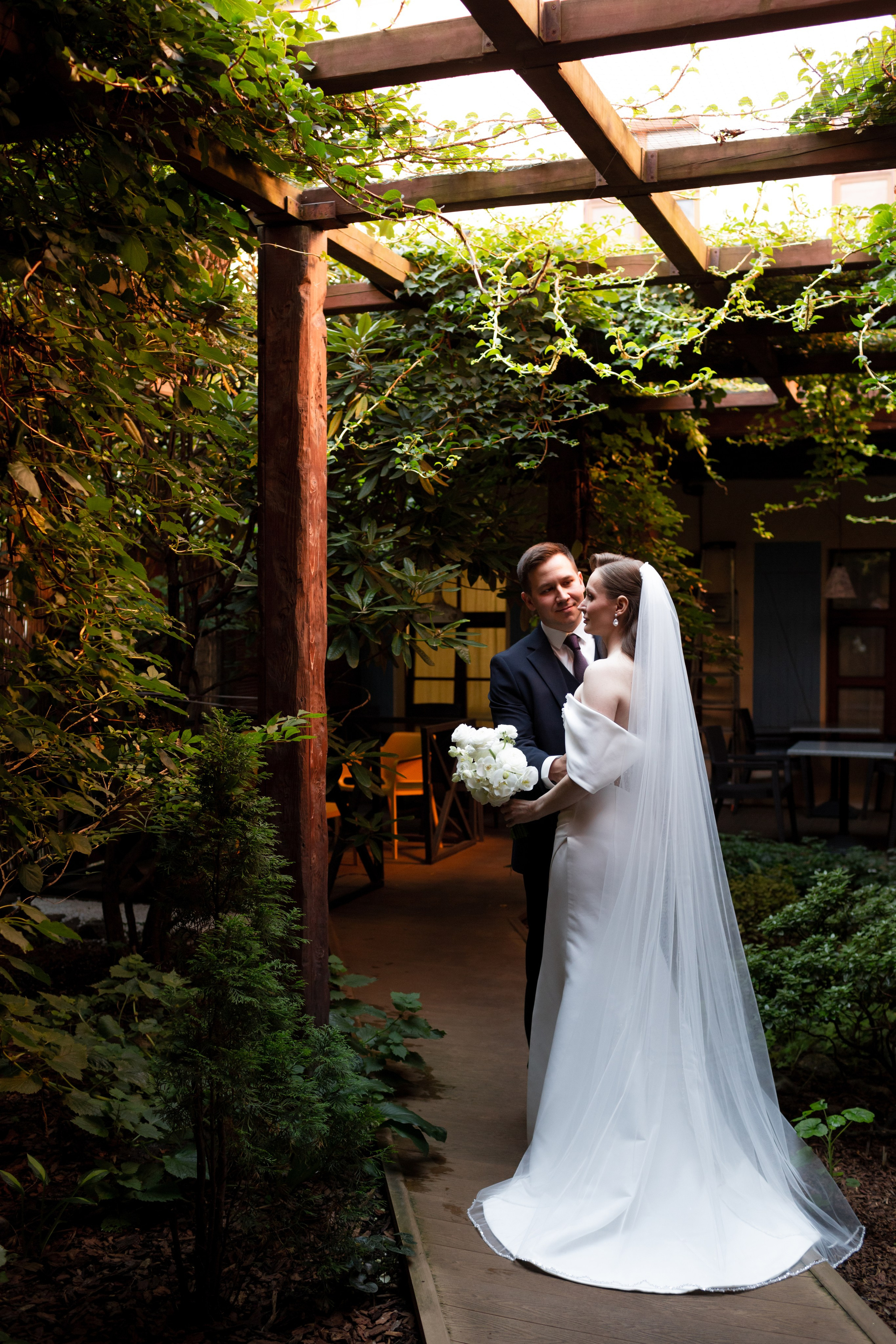 Невеста в платье с длинным шлейфом, разрезом и длинной фатой в оранжерее.