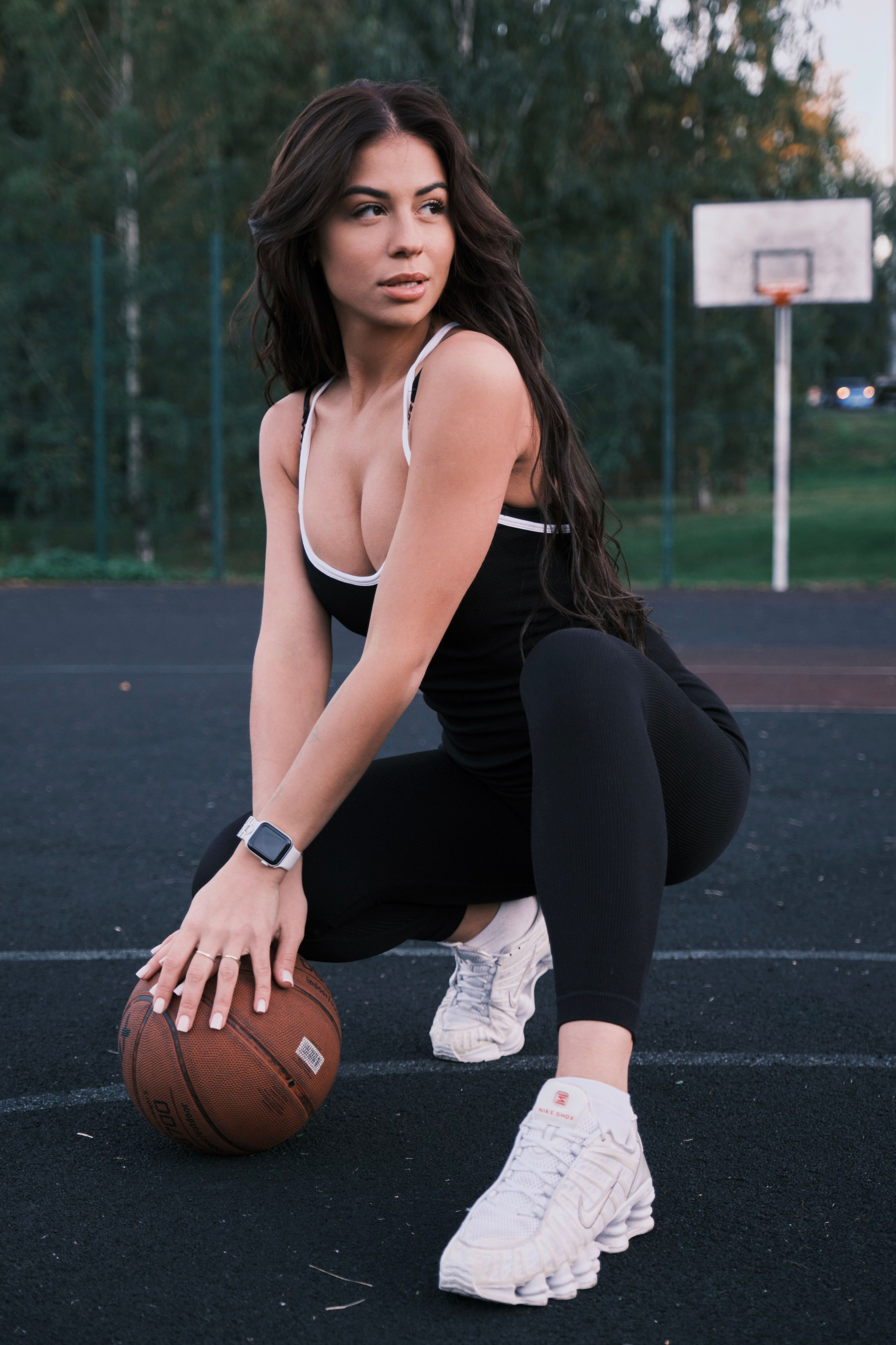 Девушка в черном спортивном костюме сидит на корточках на баскетбольной площадке, опираясь рукой на мяч.