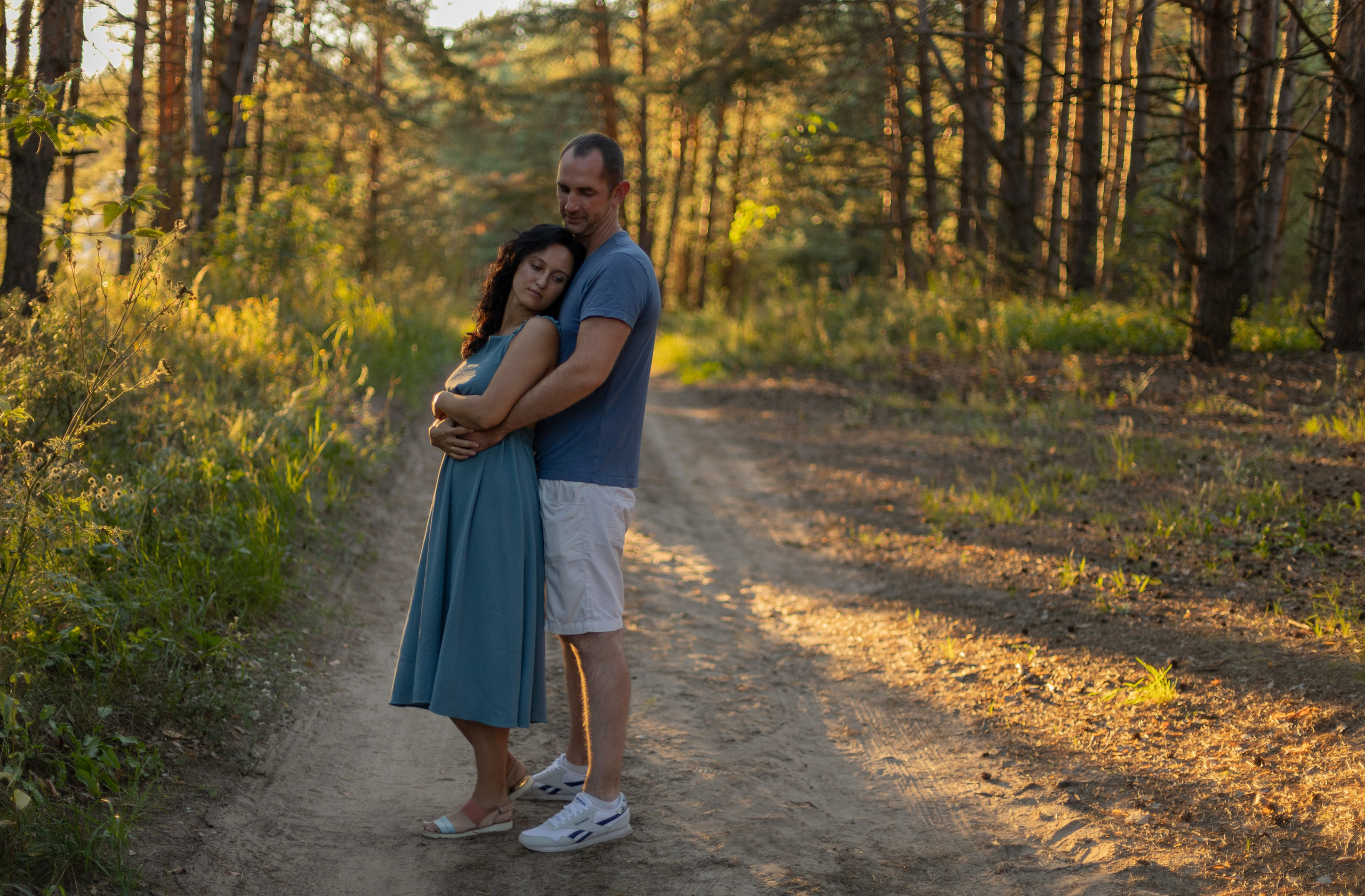 Фотосессия пары в сосновом лесу летом на закате, девушка стоит спиной к мужчине, мужчина обнимает девушку